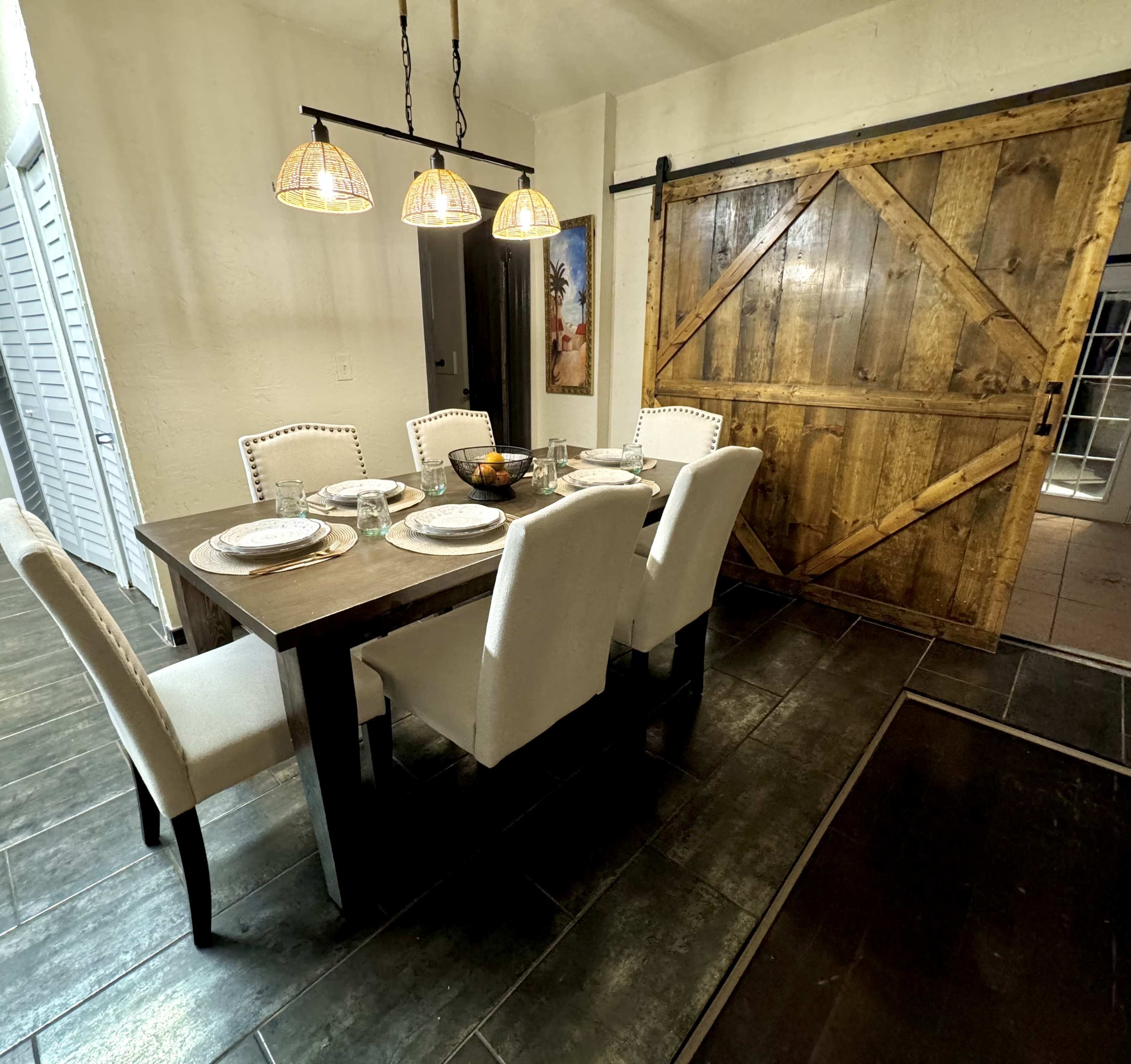 A dining room features a wooden table set for four, with white upholstered chairs, and a large sliding barn door made of wood in the background.