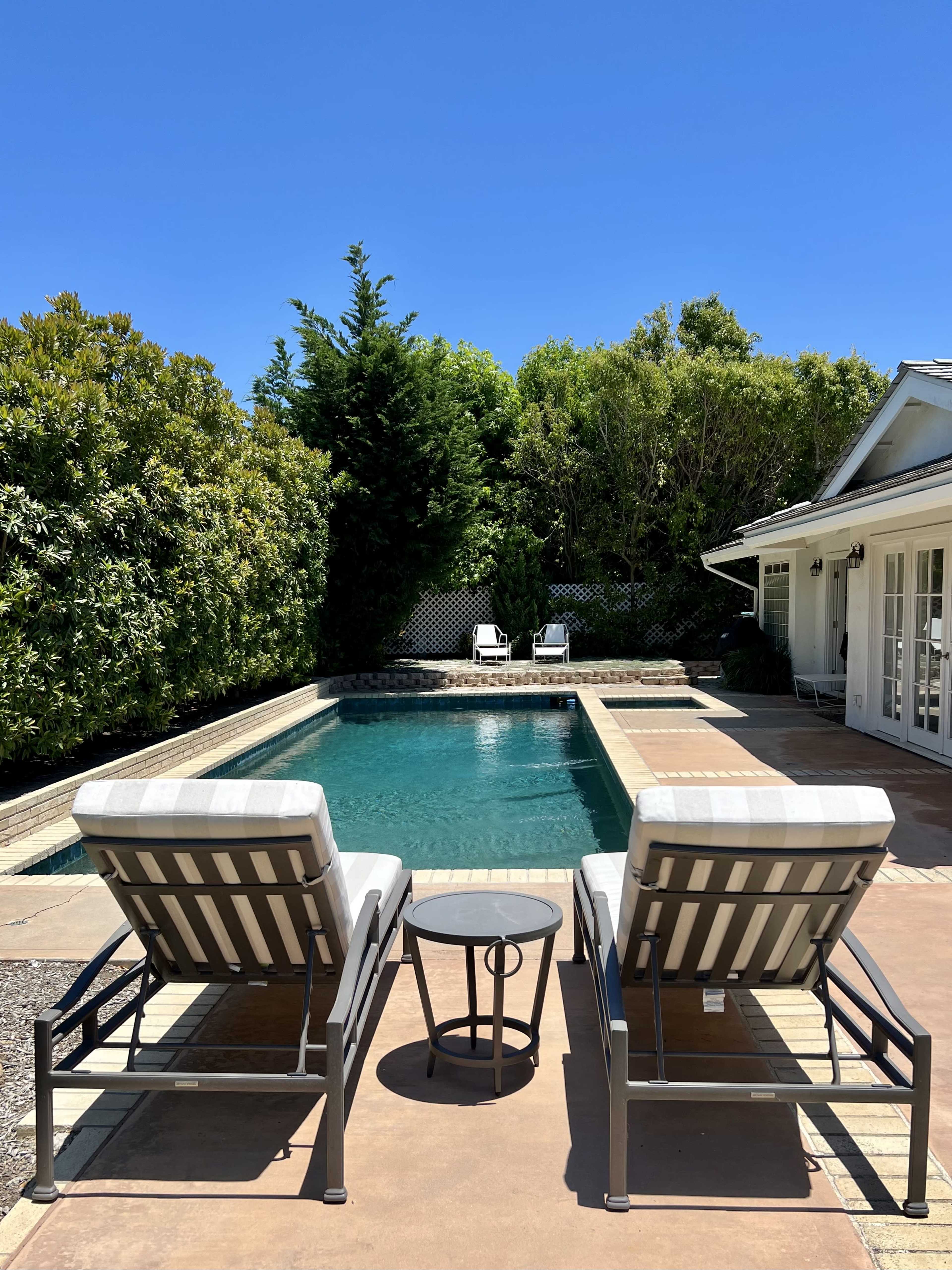 Two reclining lounge chairs flank a small table beside a swimming pool surrounded by green foliage and a sunny blue sky.