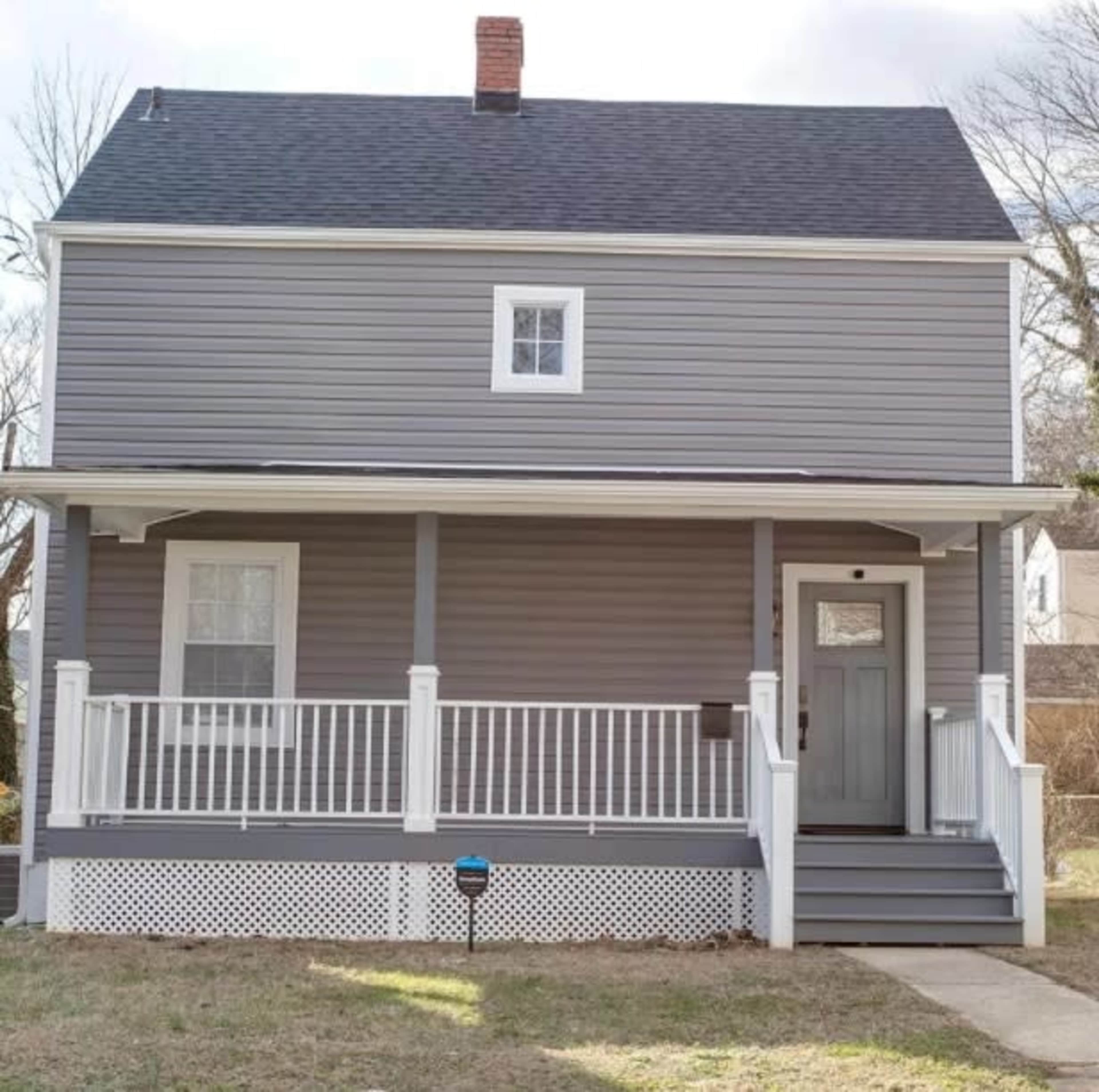 A two-story gray house with a front porch, white railings, and a single window on the second floor.