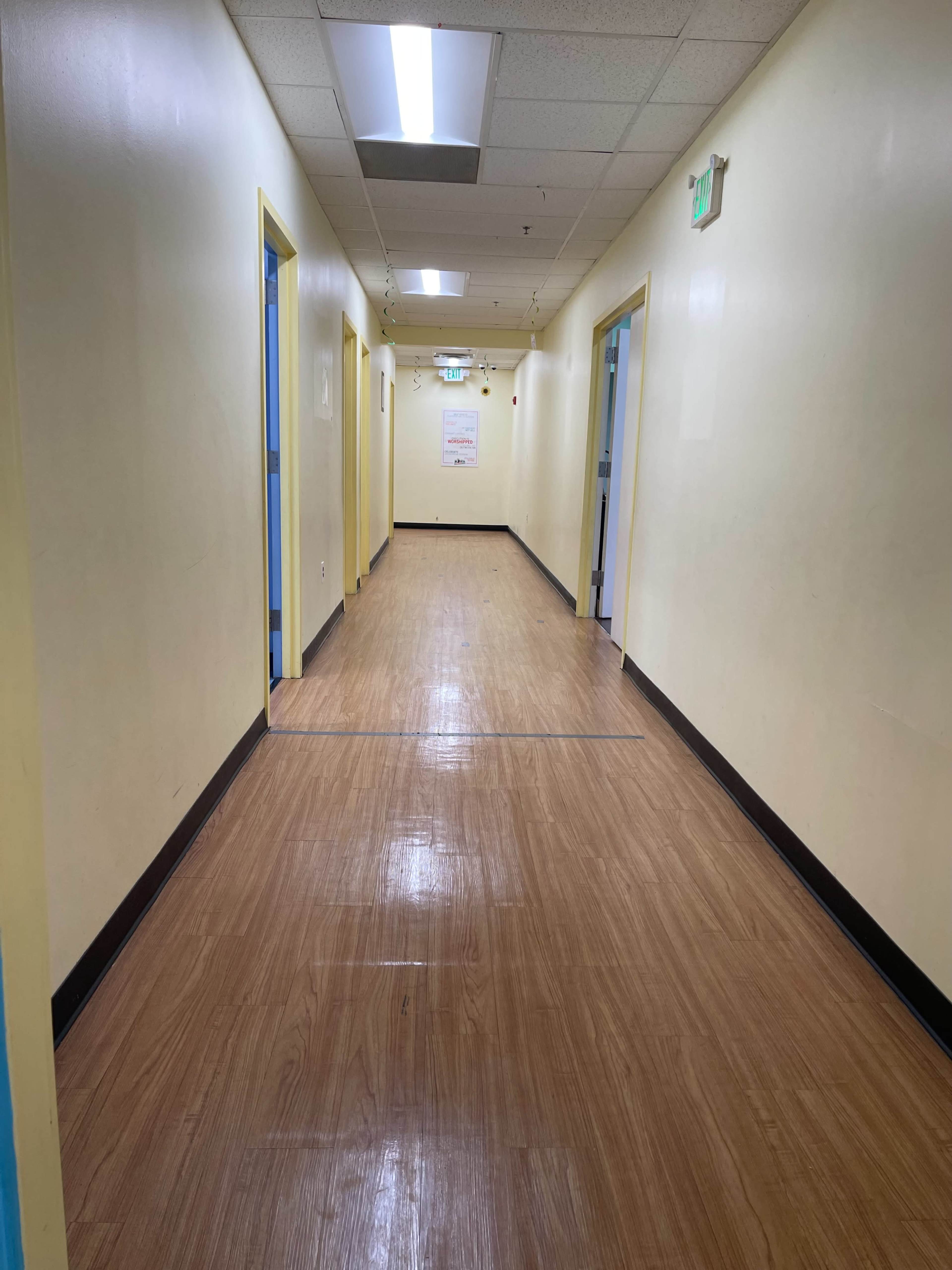 A long, brightly lit hallway with yellow walls and wooden flooring, lined with blue doorways on either side.