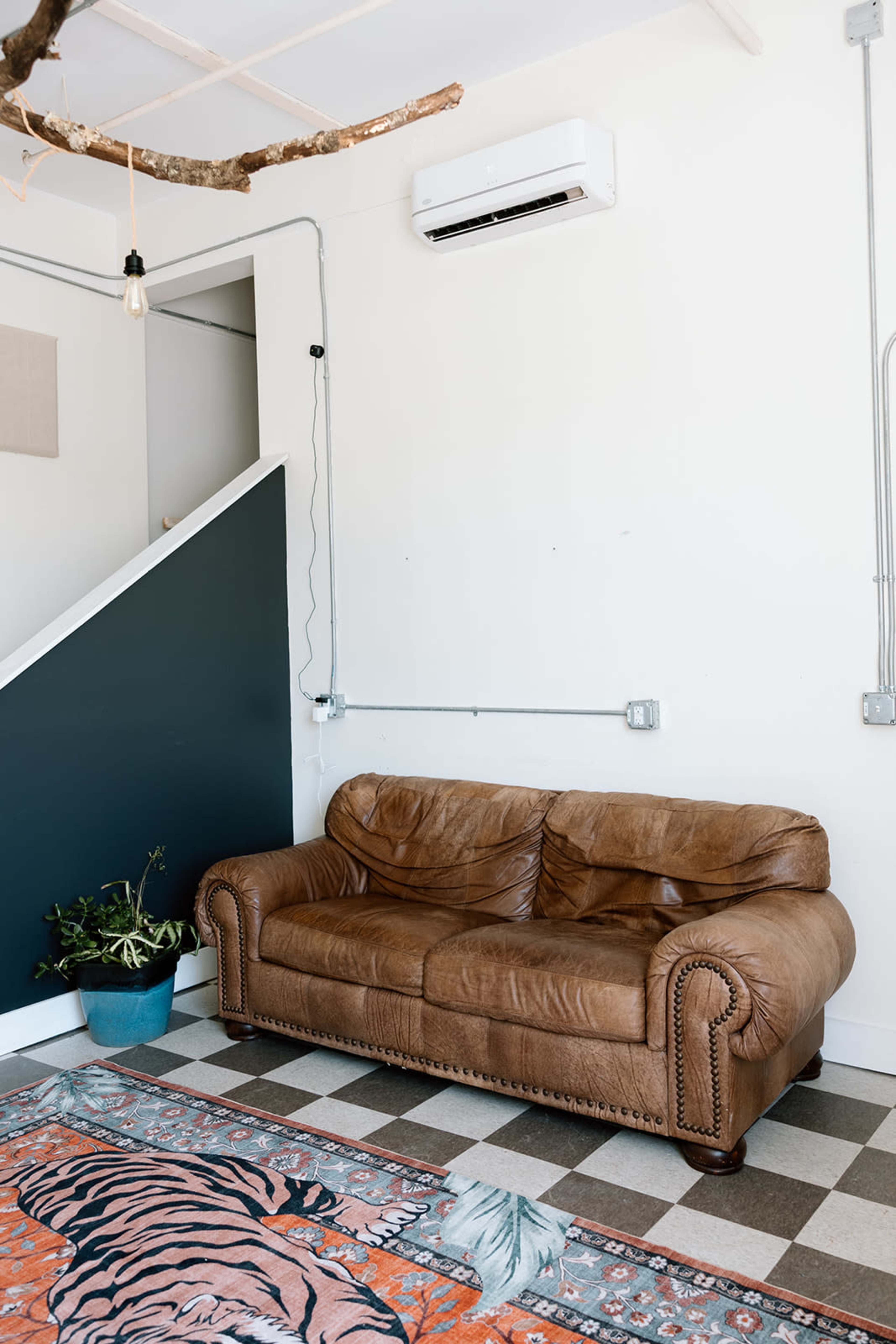 The image shows a brown leather sofa next to a potted plant on a patterned rug in a room with a staircase and wall-mounted air conditioning unit.