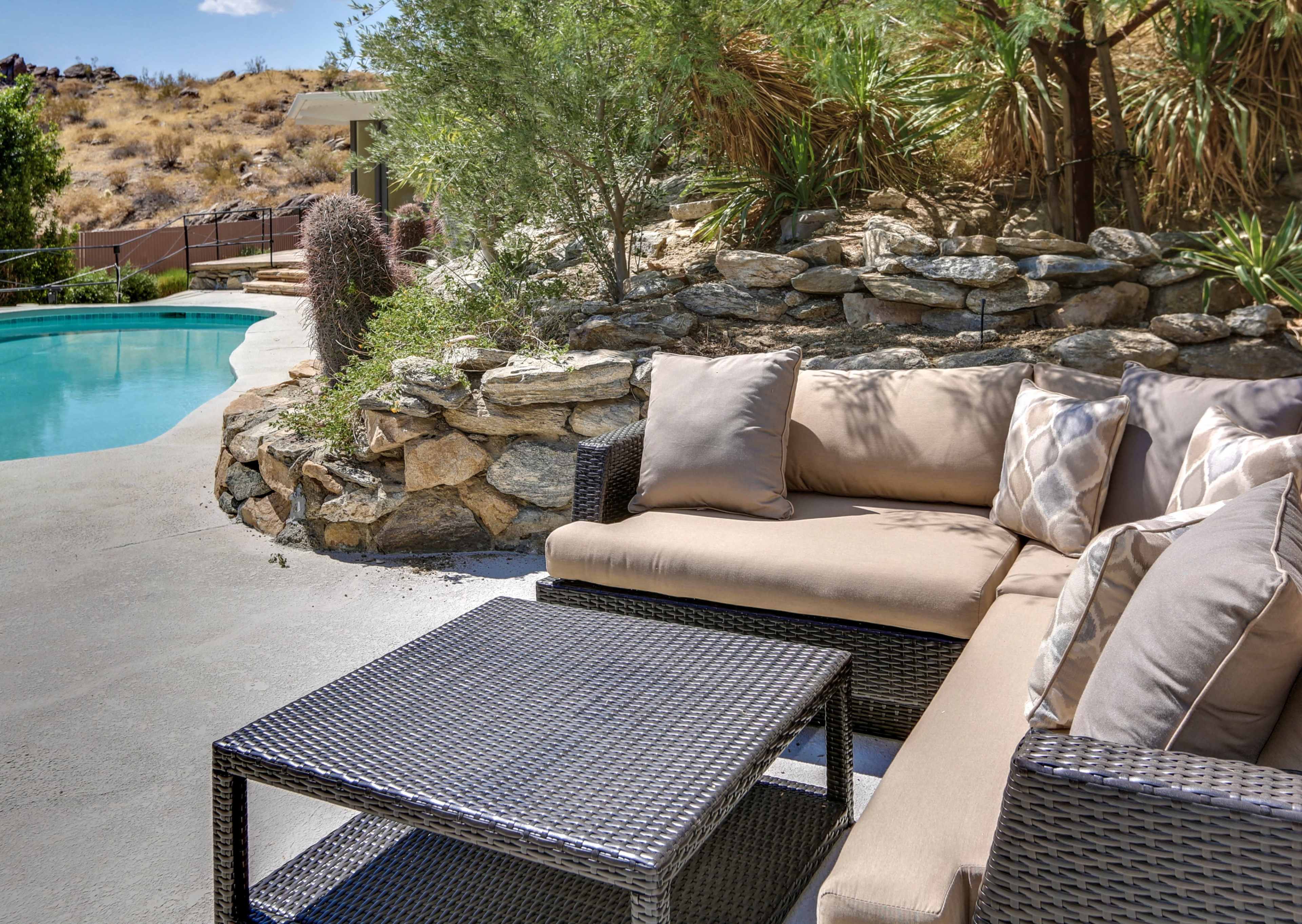 A sectional sofa and coffee table sit beside a swimming pool surrounded by rocky landscaping and desert vegetation.