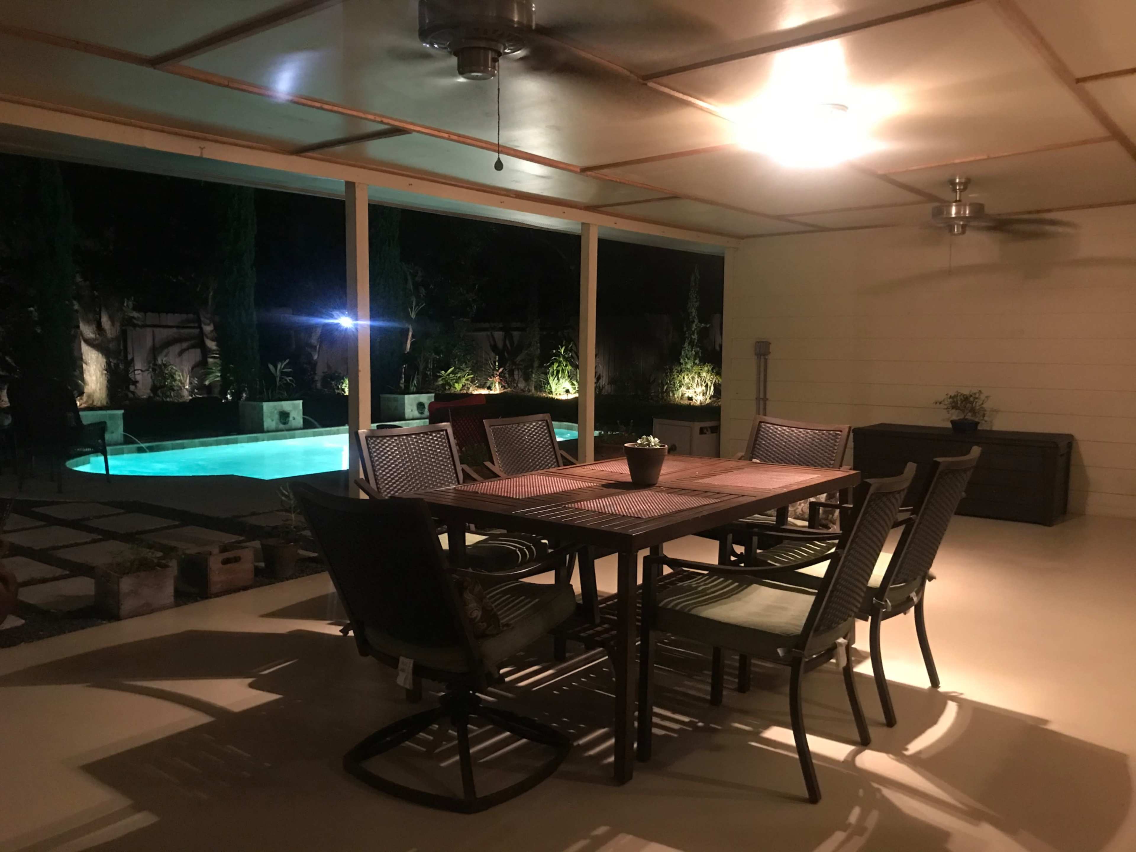 A spacious outdoor patio features a dining table surrounded by chairs, overlooking a lit swimming pool surrounded by greenery.