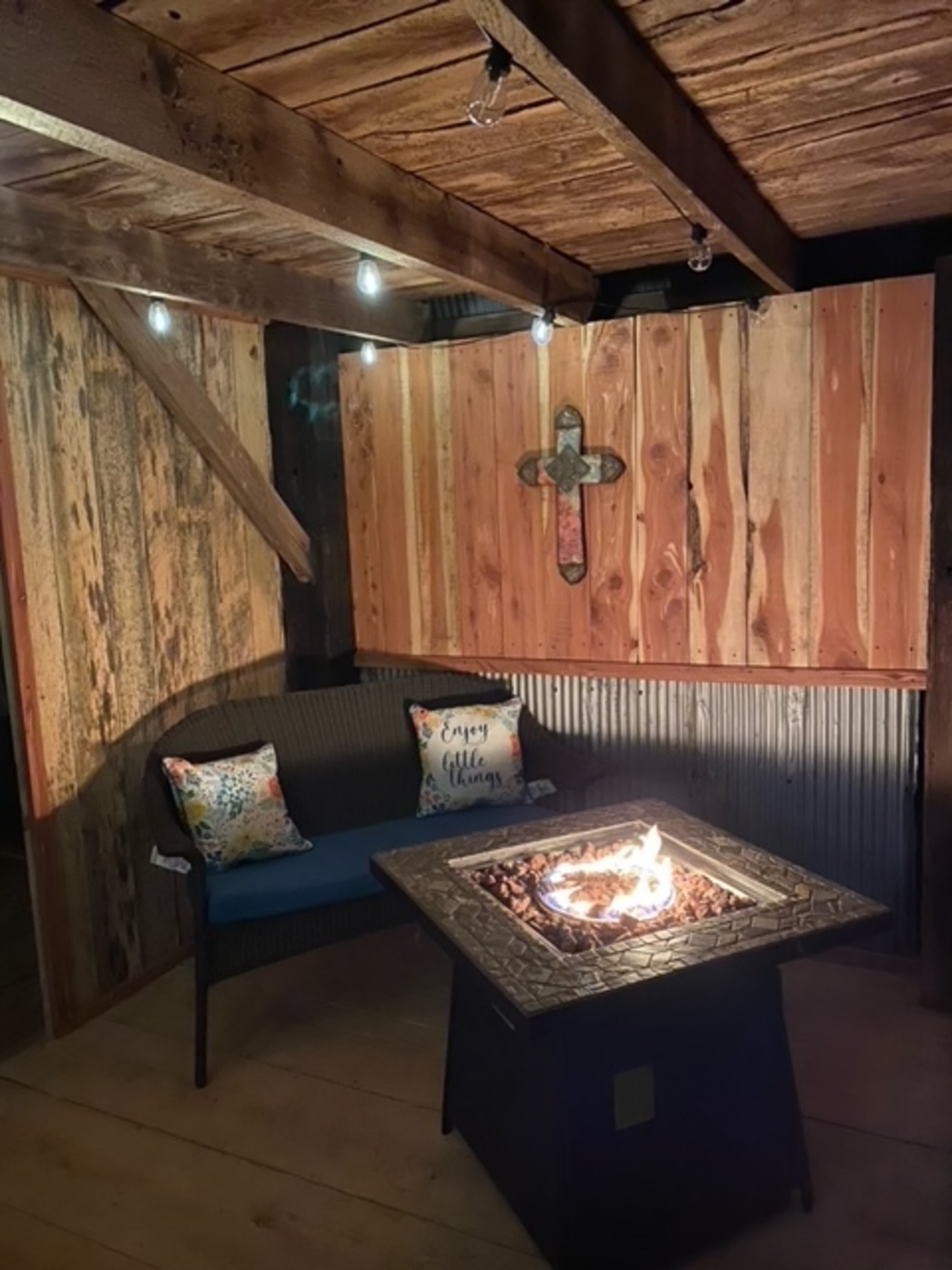 The image shows a cozy outdoor seating area featuring a fire pit surrounded by wooden walls and accent lighting.