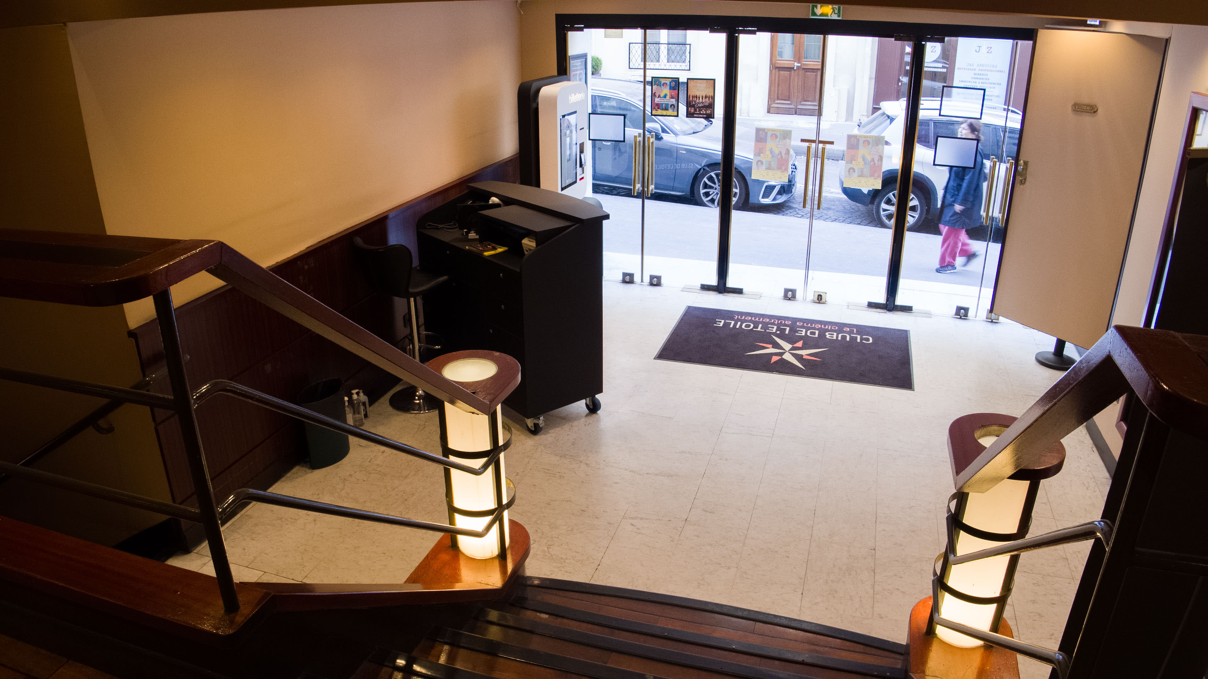 A view from the top of a staircase leading into a lobby area with a reception desk and glass doors opening to the street.