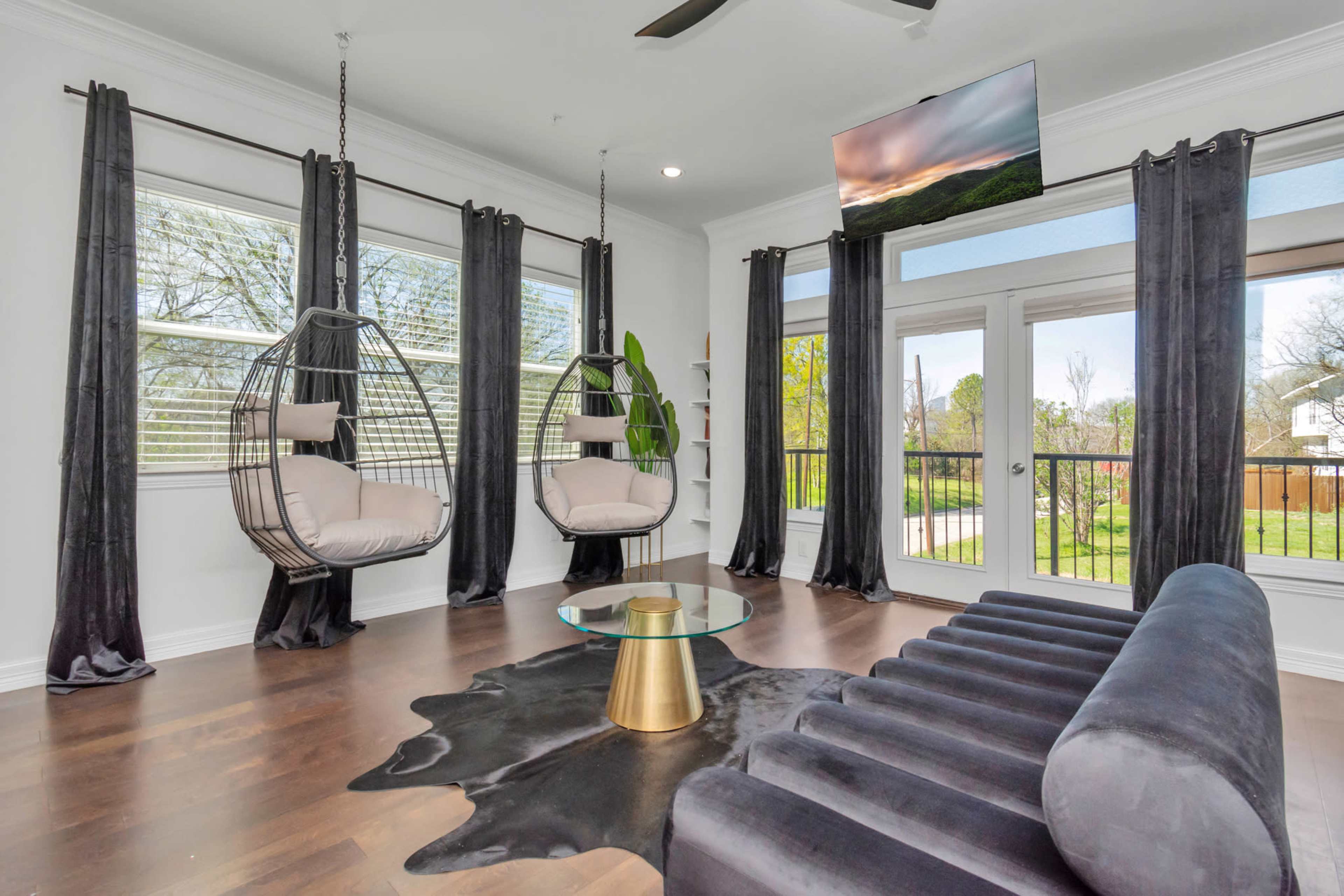 A modern living room featuring two hanging chairs, a glass coffee table, and a sleek sofa, with large windows overlooking a green outdoor space.