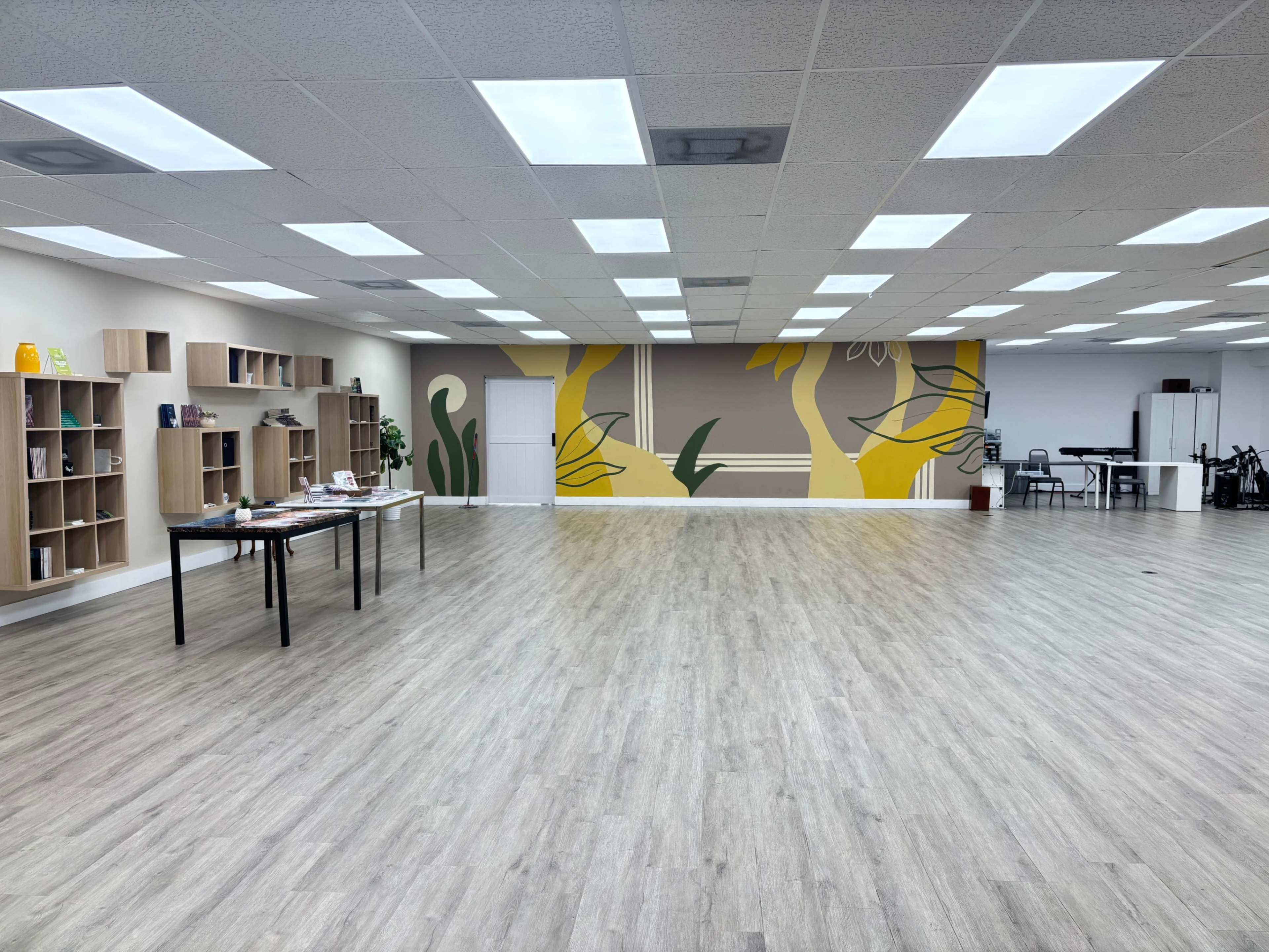 The image shows a spacious room with wooden flooring, featuring a wall of shelves on one side and a large mural with yellow designs on the opposite wall.