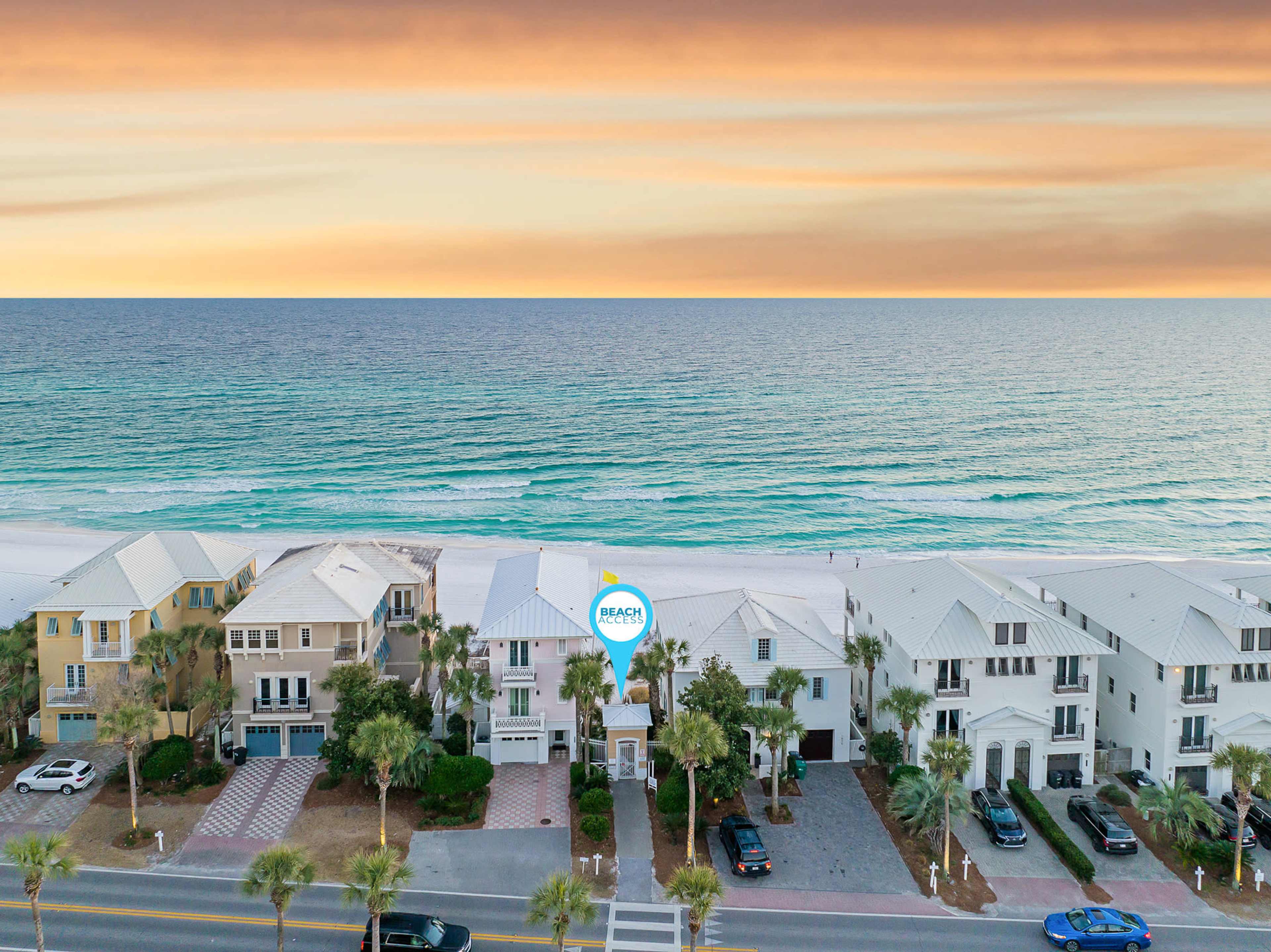 A row of beach houses along a coastal road with a view of the ocean and sunrise in the background.