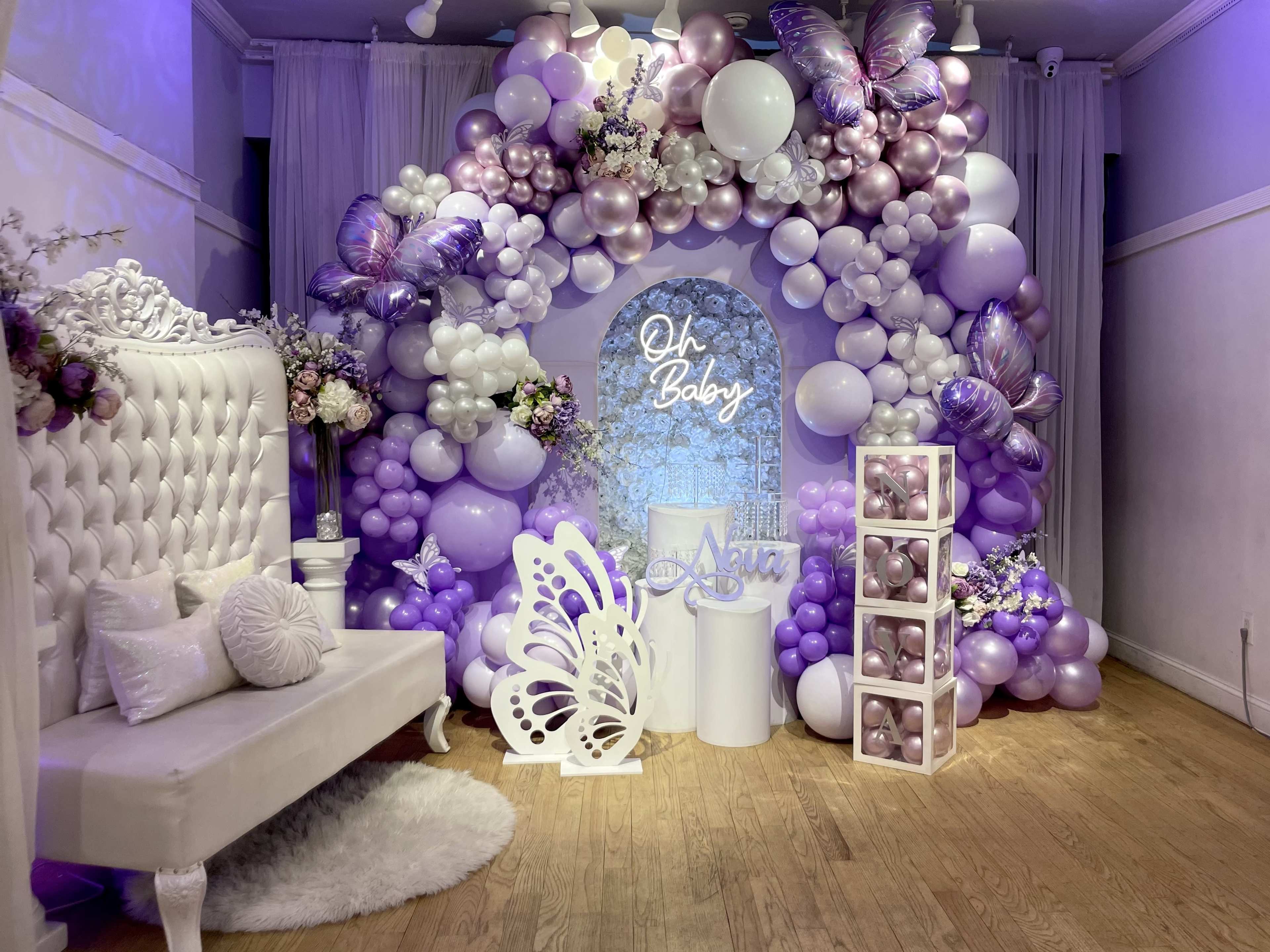 The image shows a decorative event space featuring an arch made of balloons in various shades of purple, alongside a plush white seating area and a display of gift boxes.