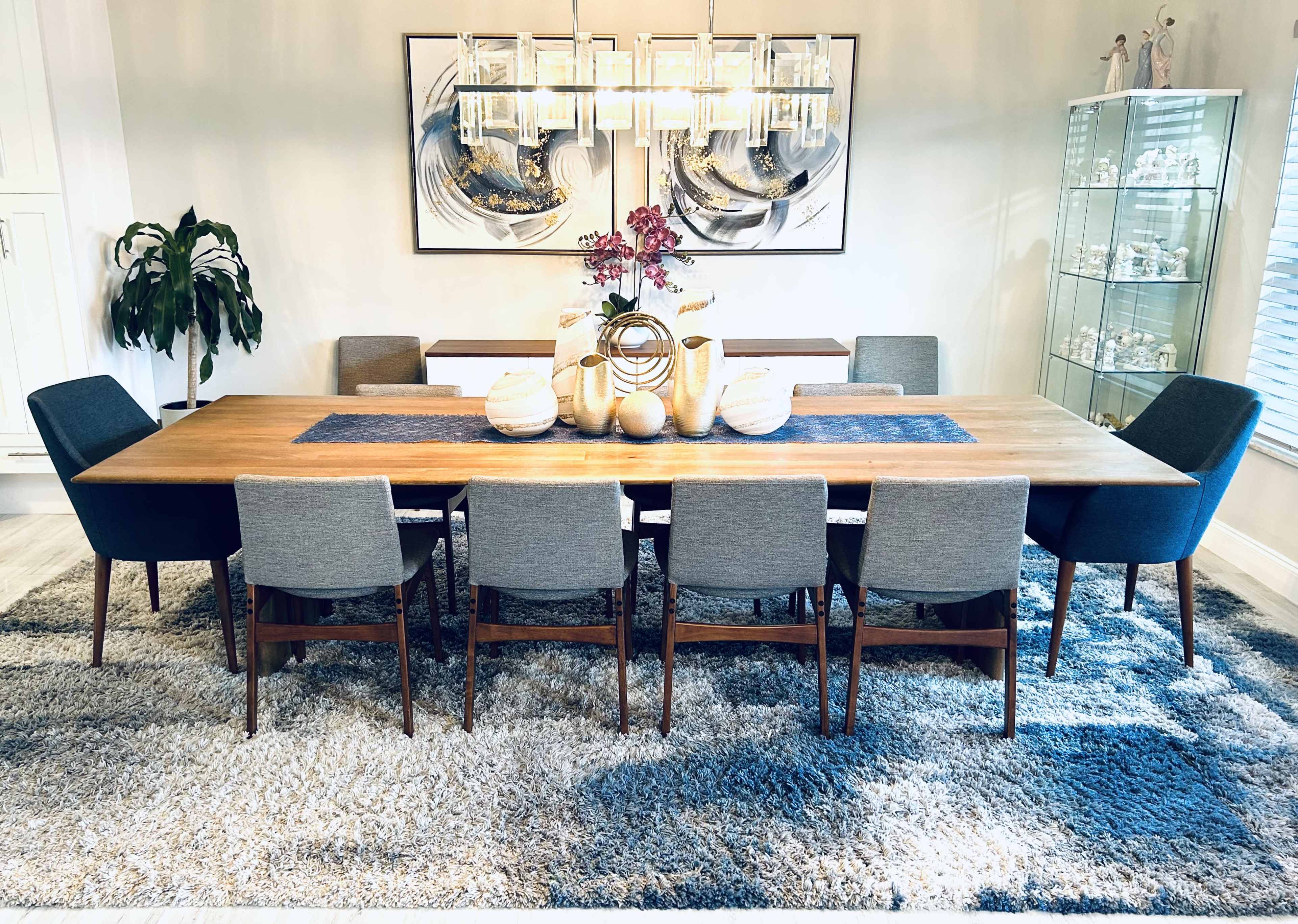 A long wooden dining table is surrounded by chairs on a plush rug, with decorative items and a light fixture above.
