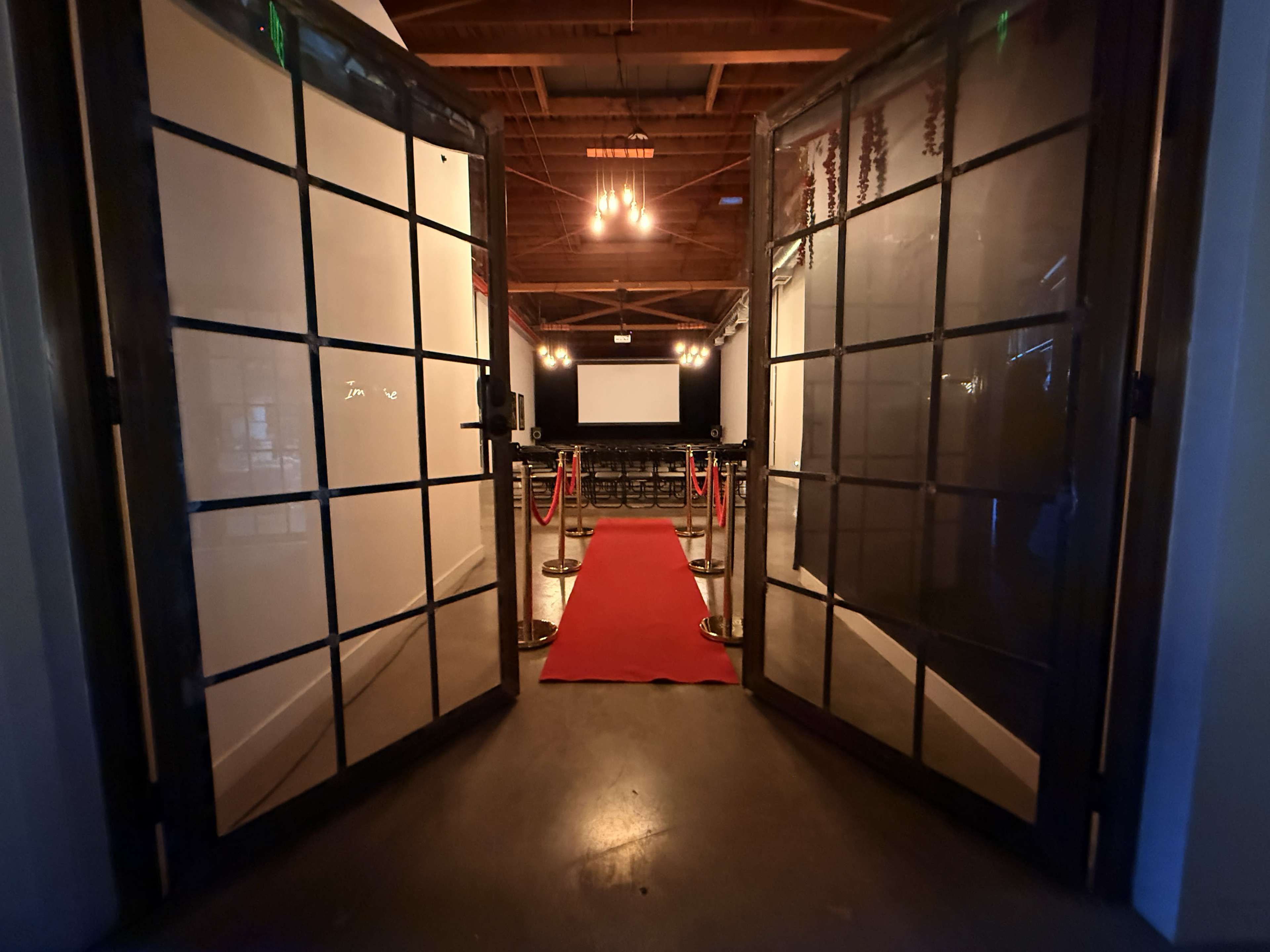 The image shows a red carpet leading into a dimly lit event space with double doors open to a projector screen and seating area in the background.