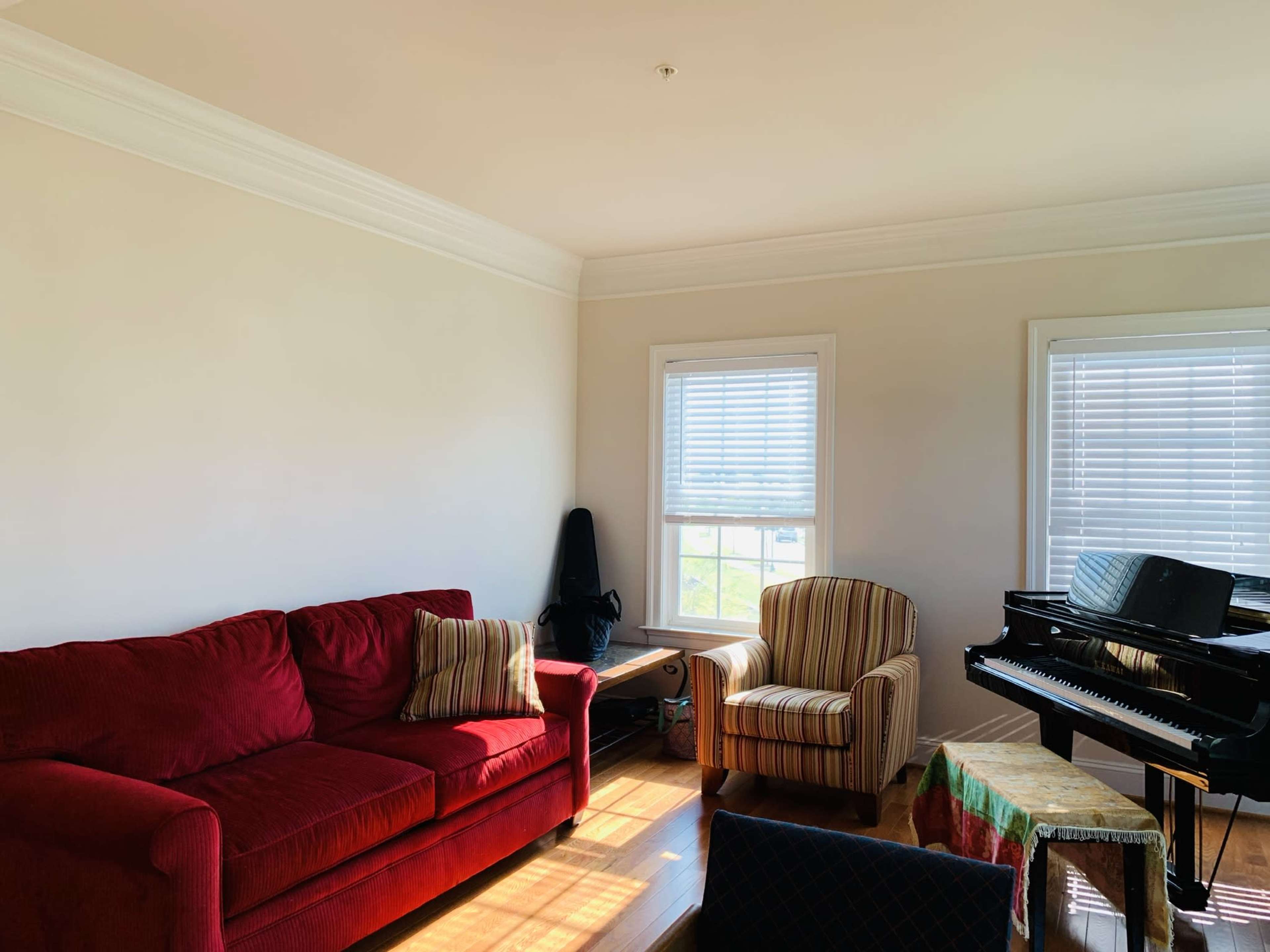 The room features a red sofa, a striped armchair, a piano, and sunlight streaming through the windows.