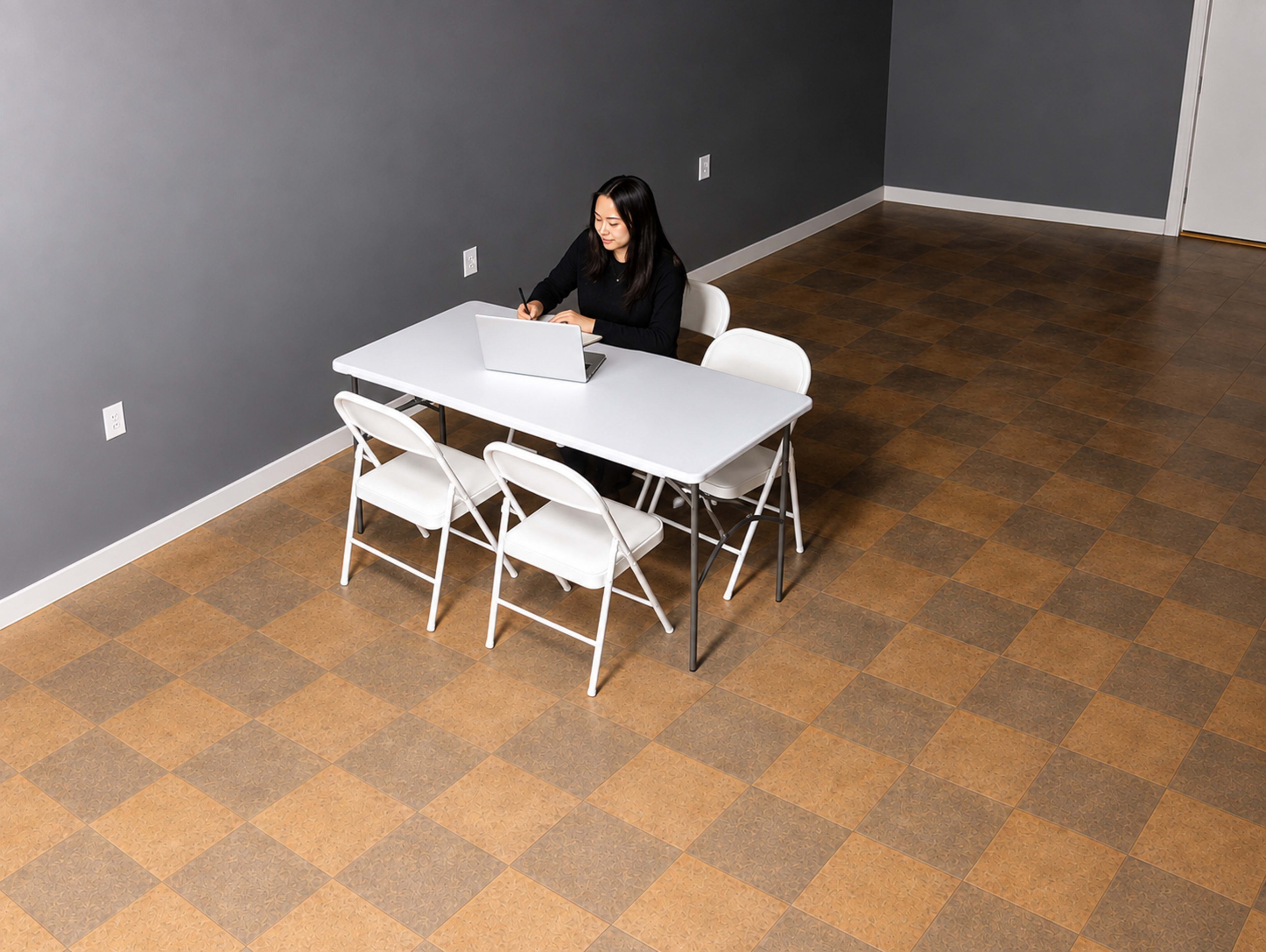 A woman is sitting at a white table with a laptop in a sparsely furnished room with a gray wall and a checkerboard floor.