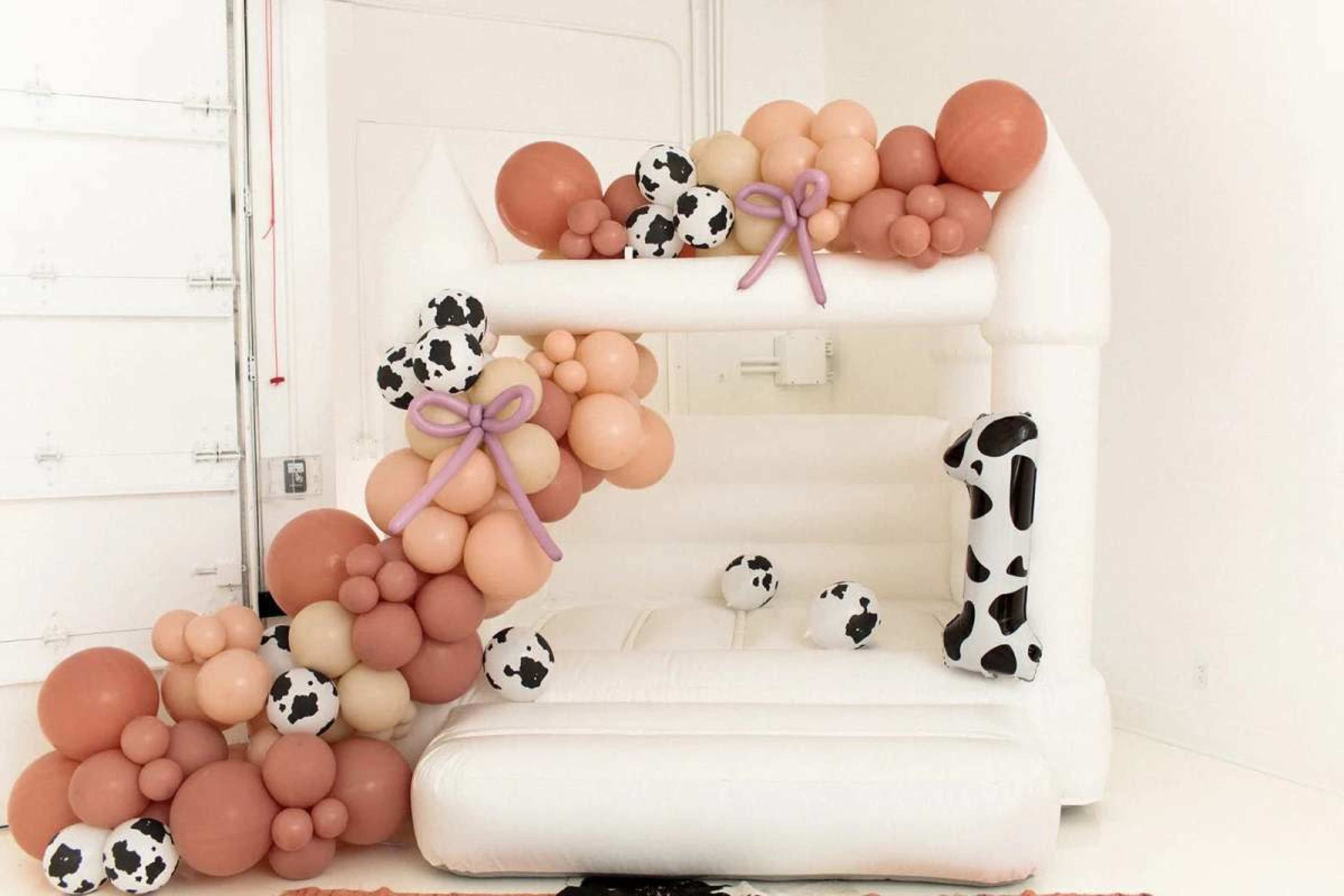 The image shows a bounce house decorated with an arrangement of balloons in various colors, including peach, cream, and black-and-white cow patterns.