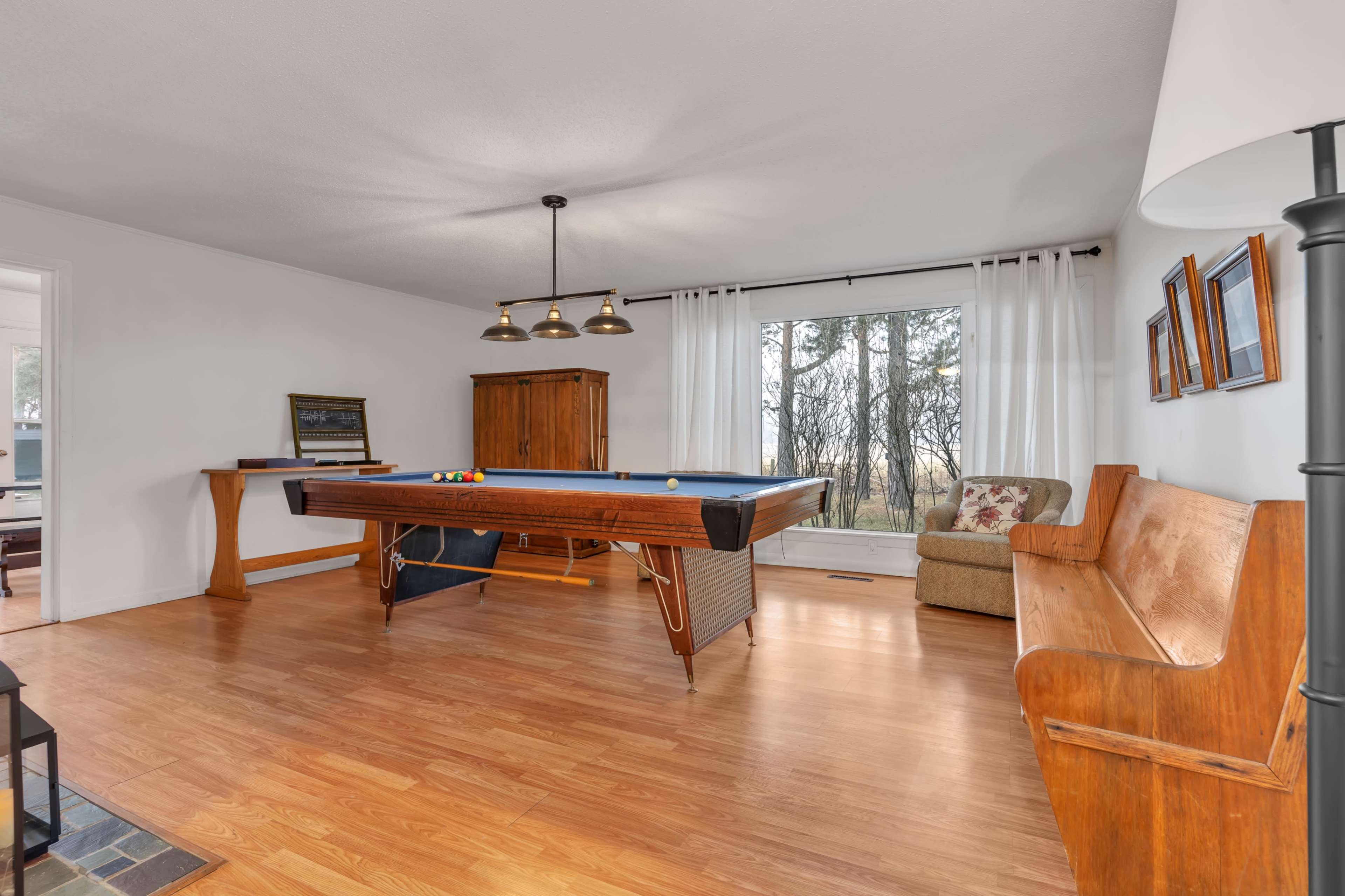 The image shows a spacious room featuring a billiard table, a wooden bench, and large windows with a view of trees outside.