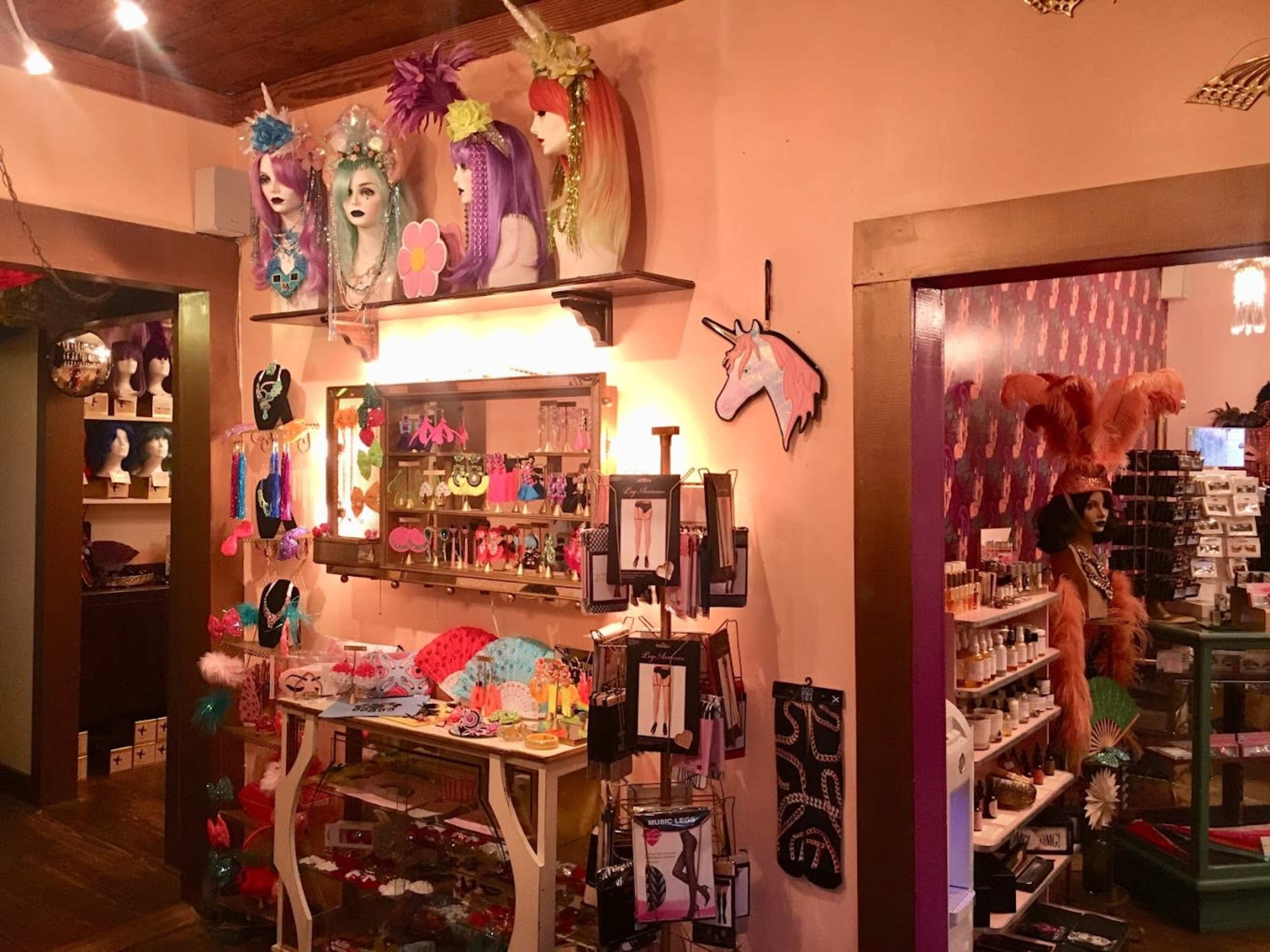 The image shows a colorful boutique filled with wigs, accessories, and a variety of displayed items against a pink wall.