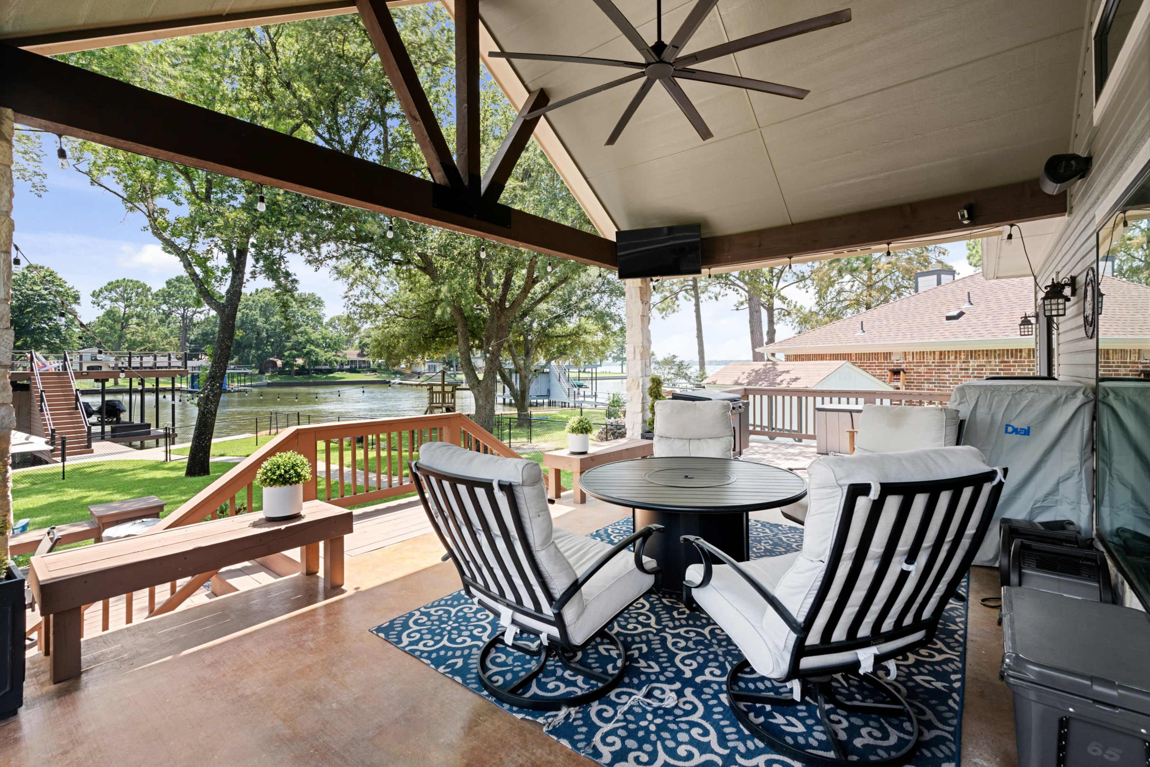 The image shows a covered outdoor seating area with two lounge chairs and a table, overlooking a serene waterfront view surrounded by trees.