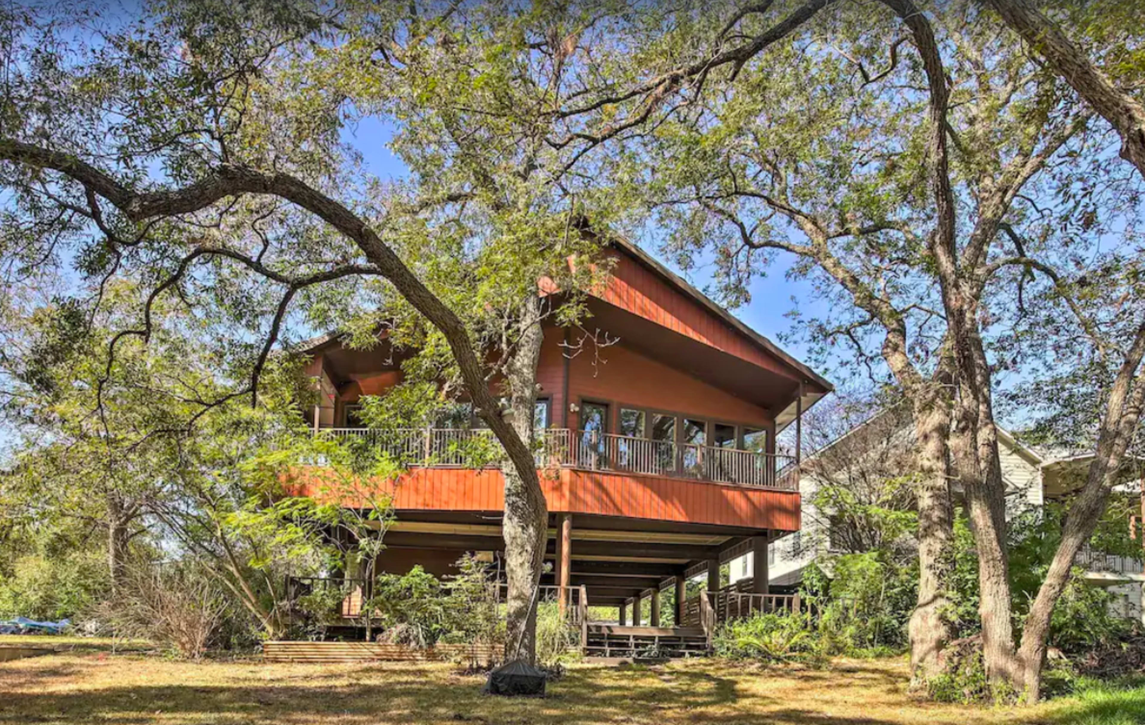 A wooden house with a large deck is elevated on stilts amid a landscape of trees and grass.