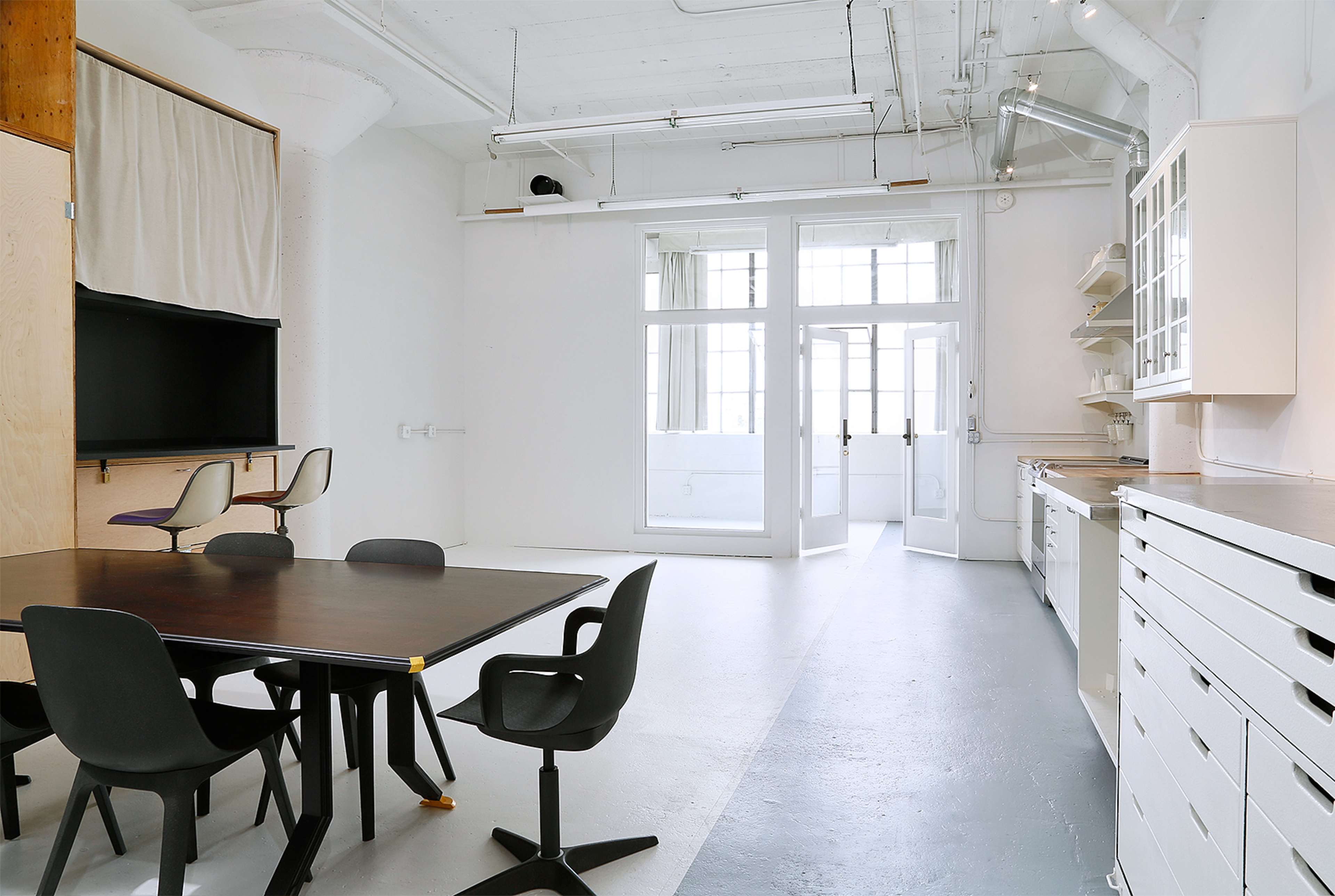 The image shows a spacious, minimalist room with a table and chairs, a wall-mounted screen, and a kitchenette area, featuring large windows that allow natural light to enter.