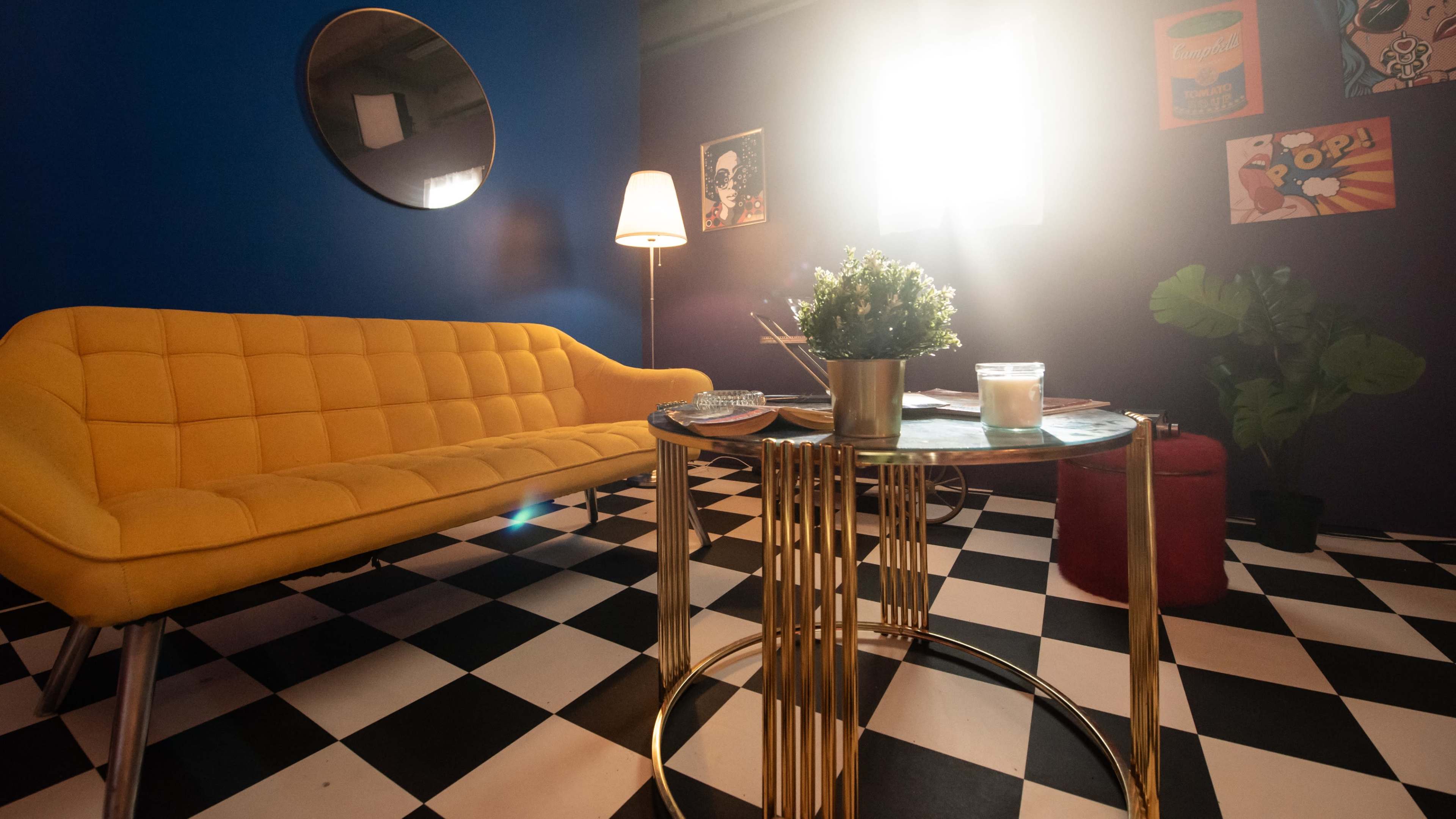 A brightly lit room features a yellow sofa, a circular mirror, a coffee table with a plant and candle, and a checkered floor.