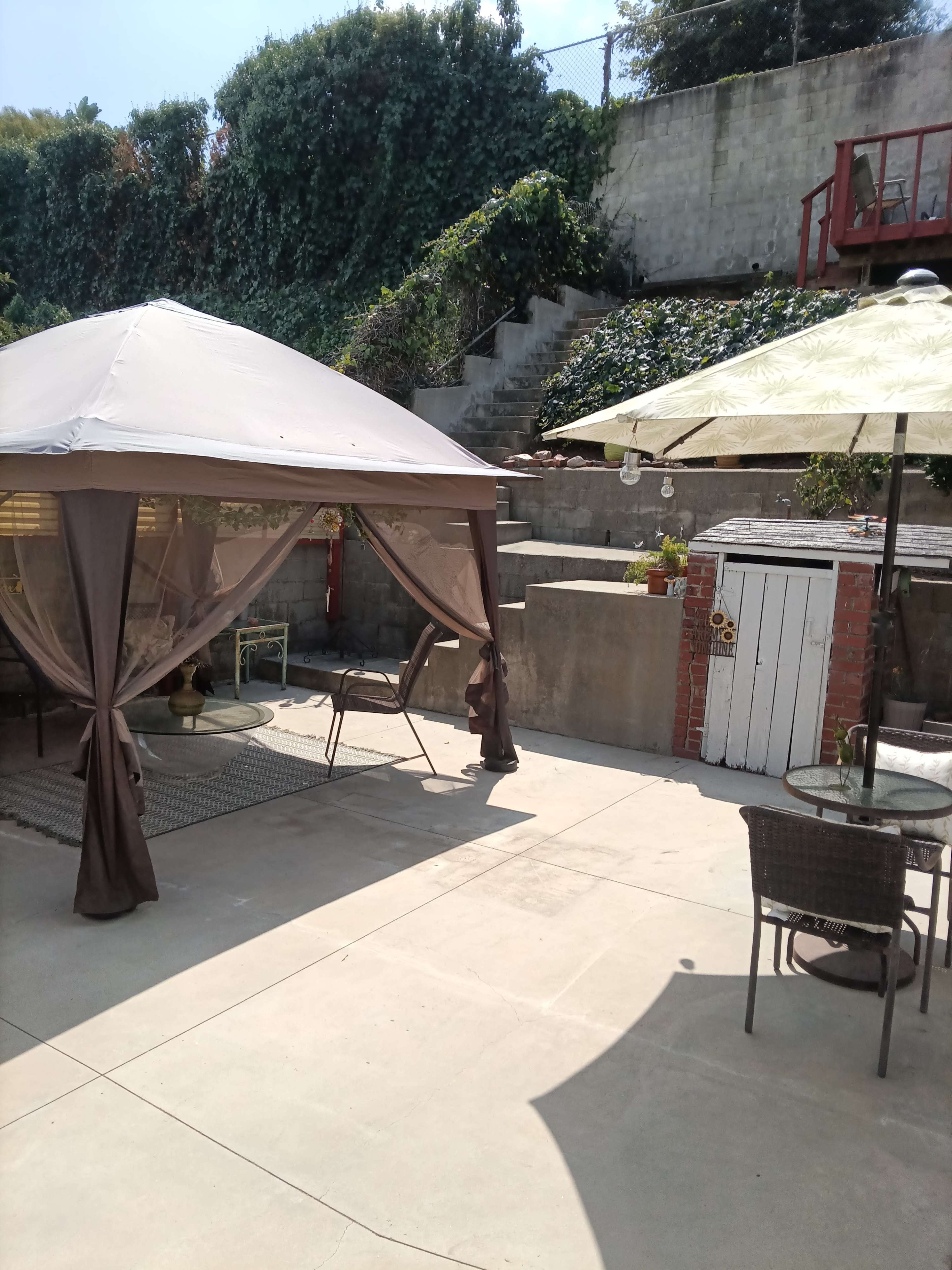 The image shows a patio area with a gazebo, seating arrangements, and a pathway leading to stairs surrounded by greenery.
