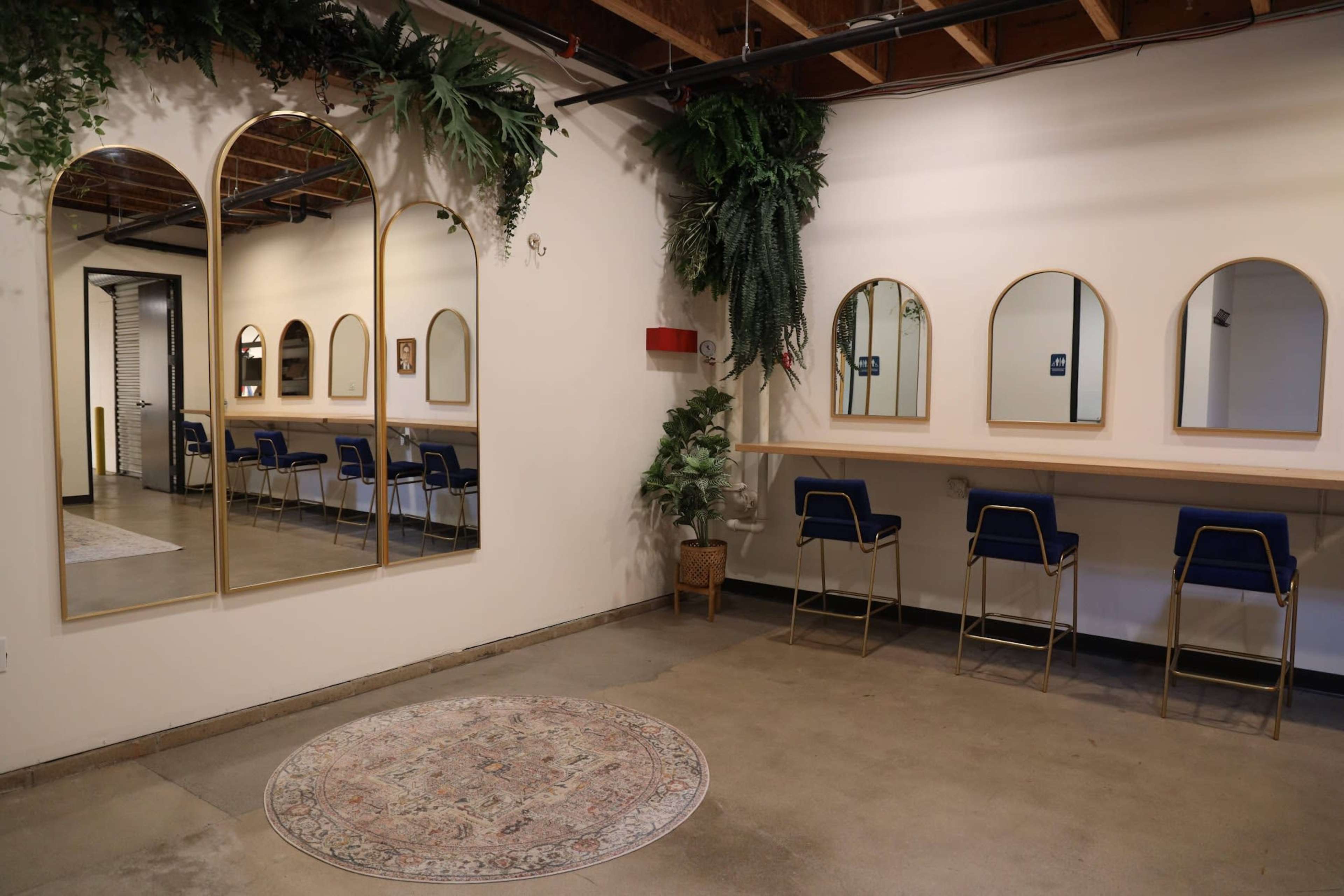 The image shows a spacious room with arched mirrors and seating areas, decorated with greenery and a patterned rug on the concrete floor.