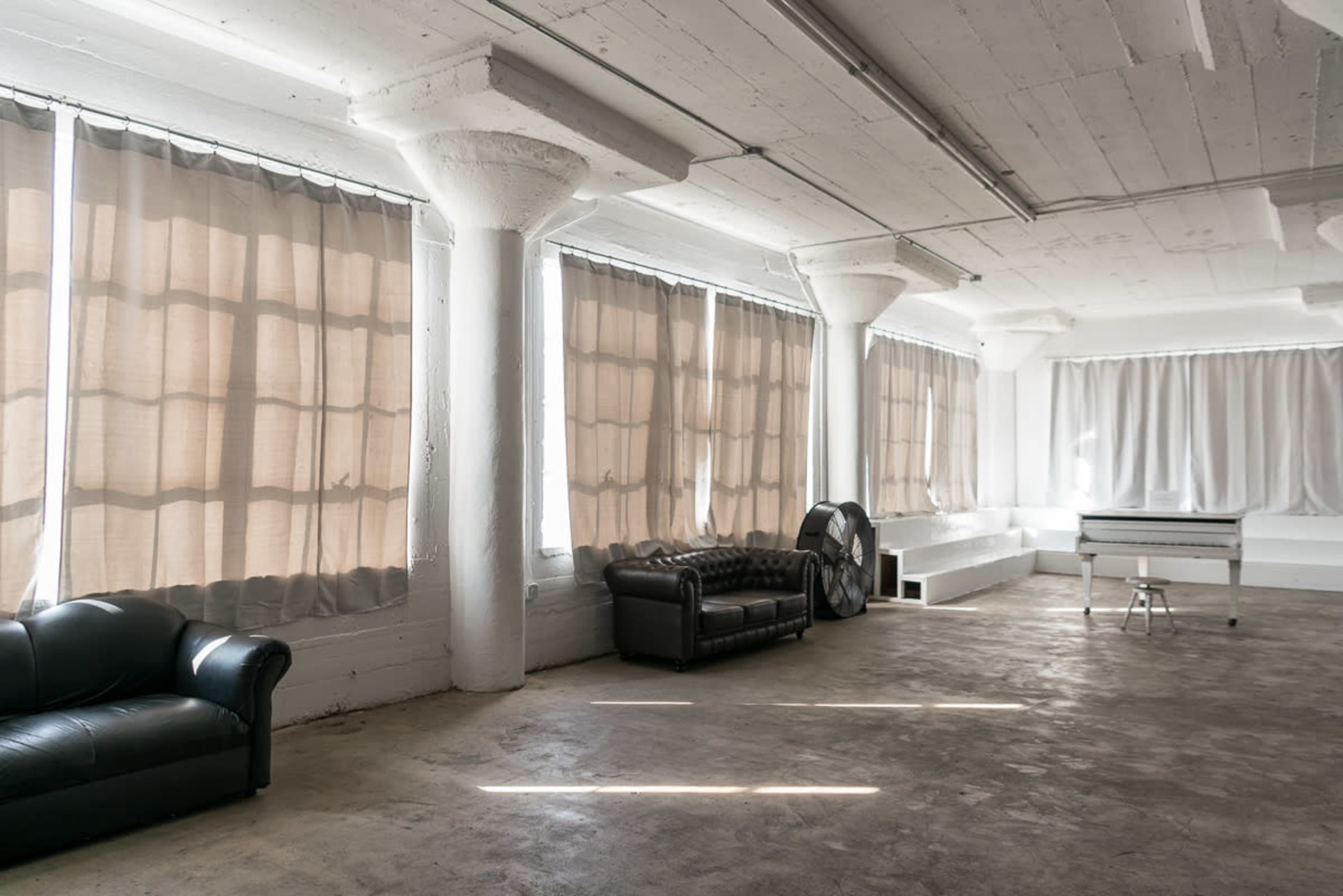 The image shows a spacious, empty room with tall windows covered by sheer curtains, featuring a leather couch and a white table.