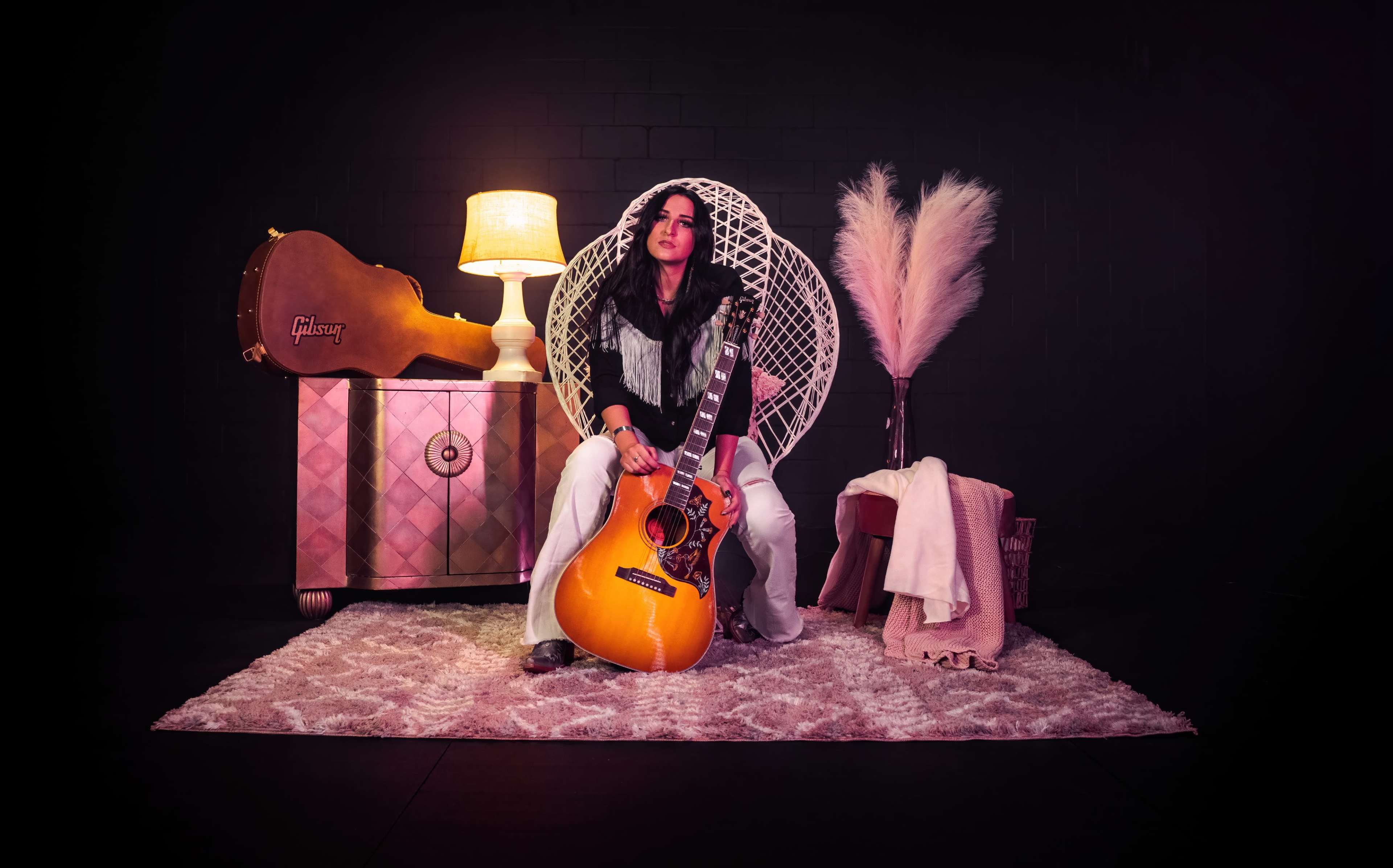 A woman sits on a chair holding a guitar, with a lamp, a decorative plant, and a suitcase in the background on a rug.