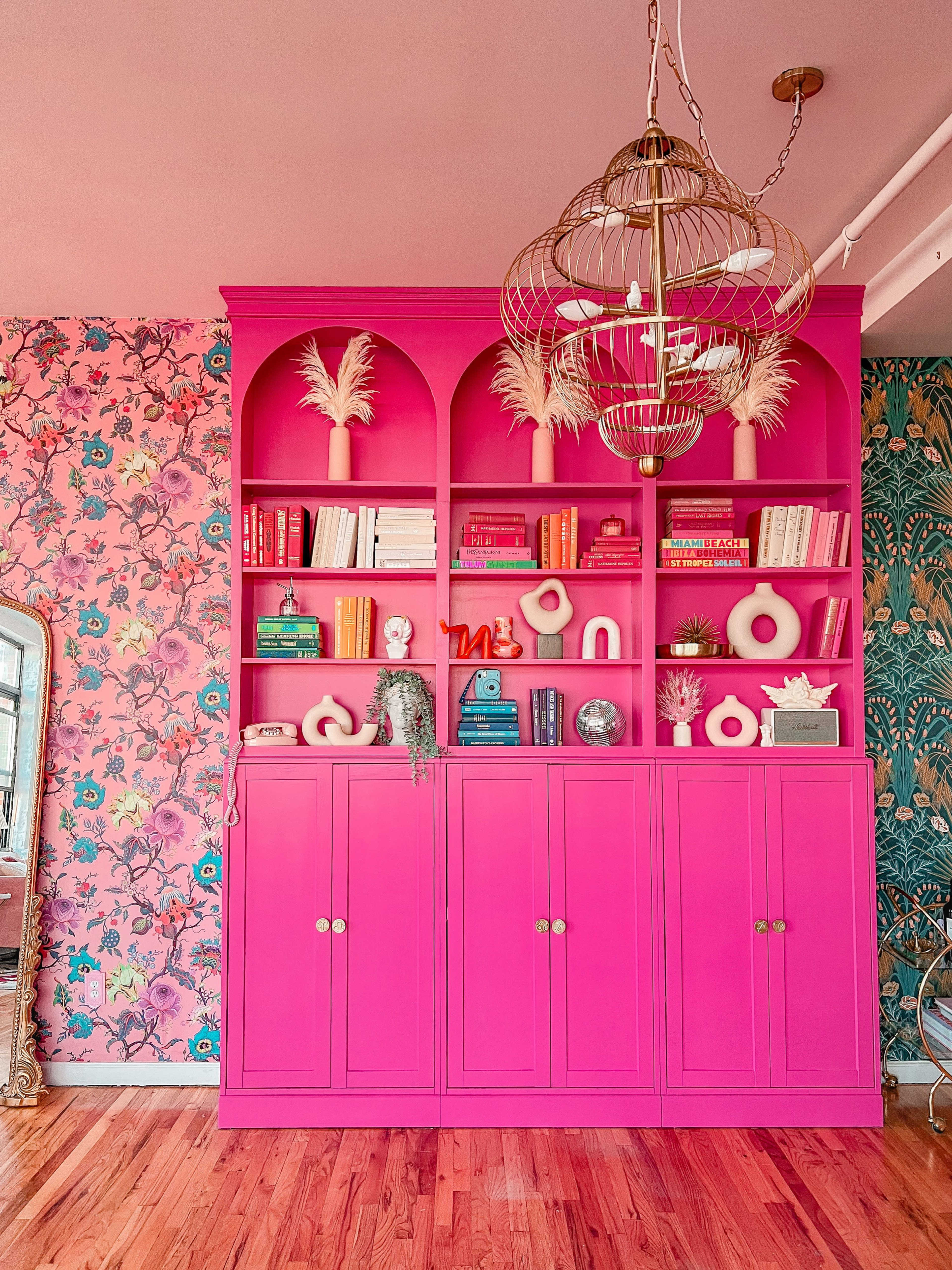 The image shows a vibrant pink bookshelf filled with colorful books and decorative items against a floral wallpaper background.