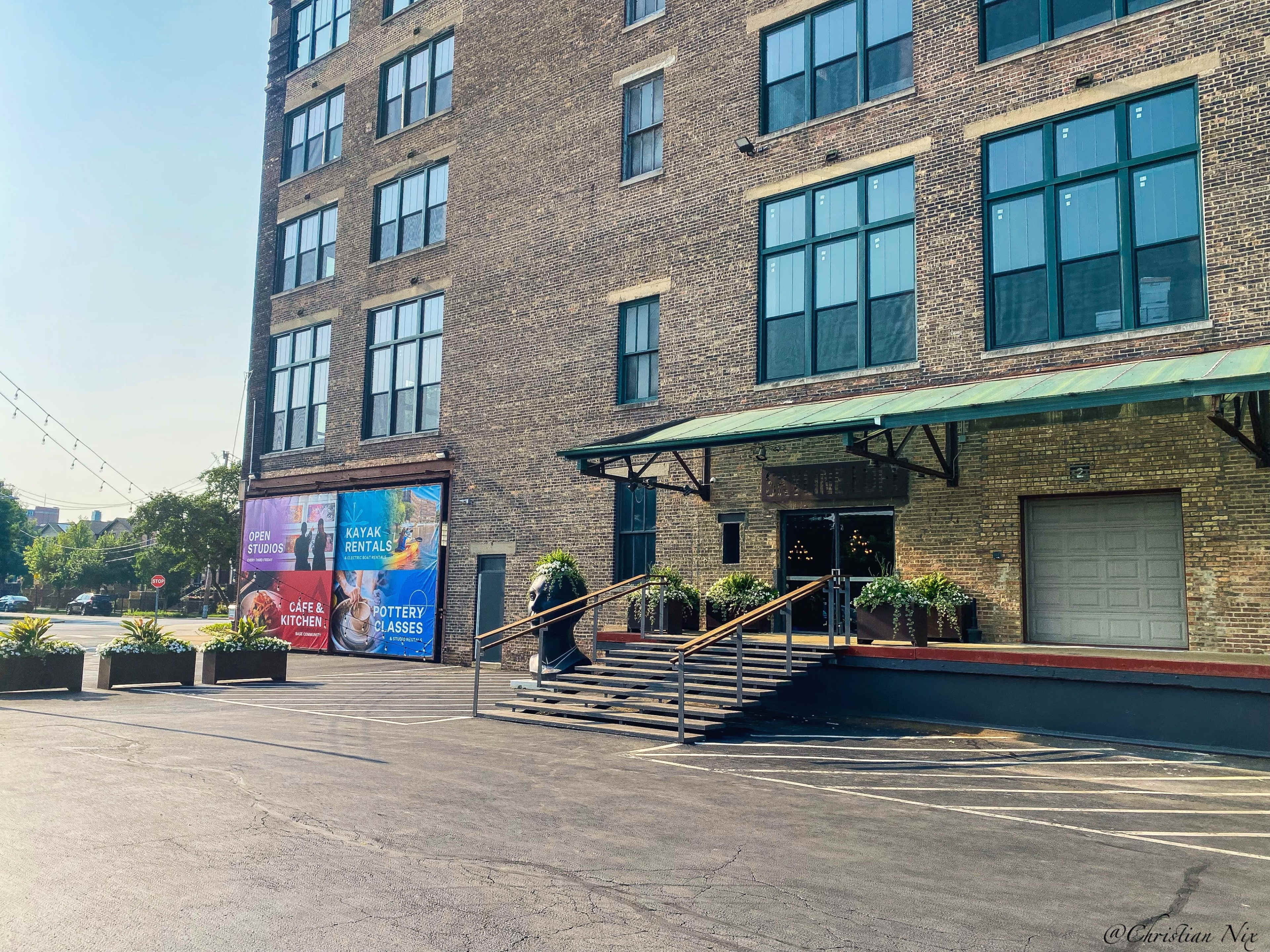 The image features a brick building with large windows and a set of steps leading to an entrance, alongside banners advertising various events and services.