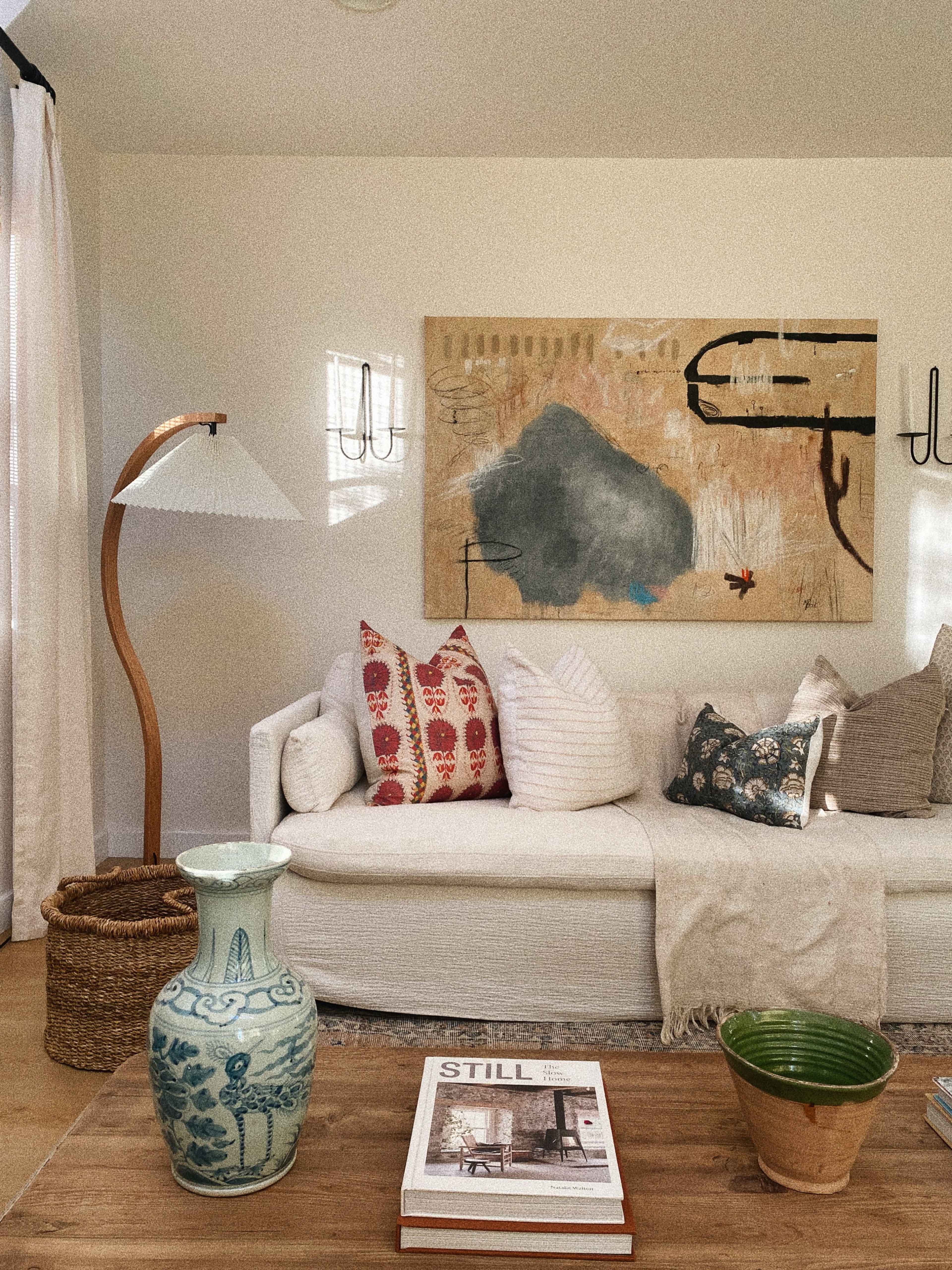 A cozy living room features a white sofa adorned with patterned pillows, a wooden coffee table with books and decorative items, and a floor lamp beside a colorful artwork on the wall.