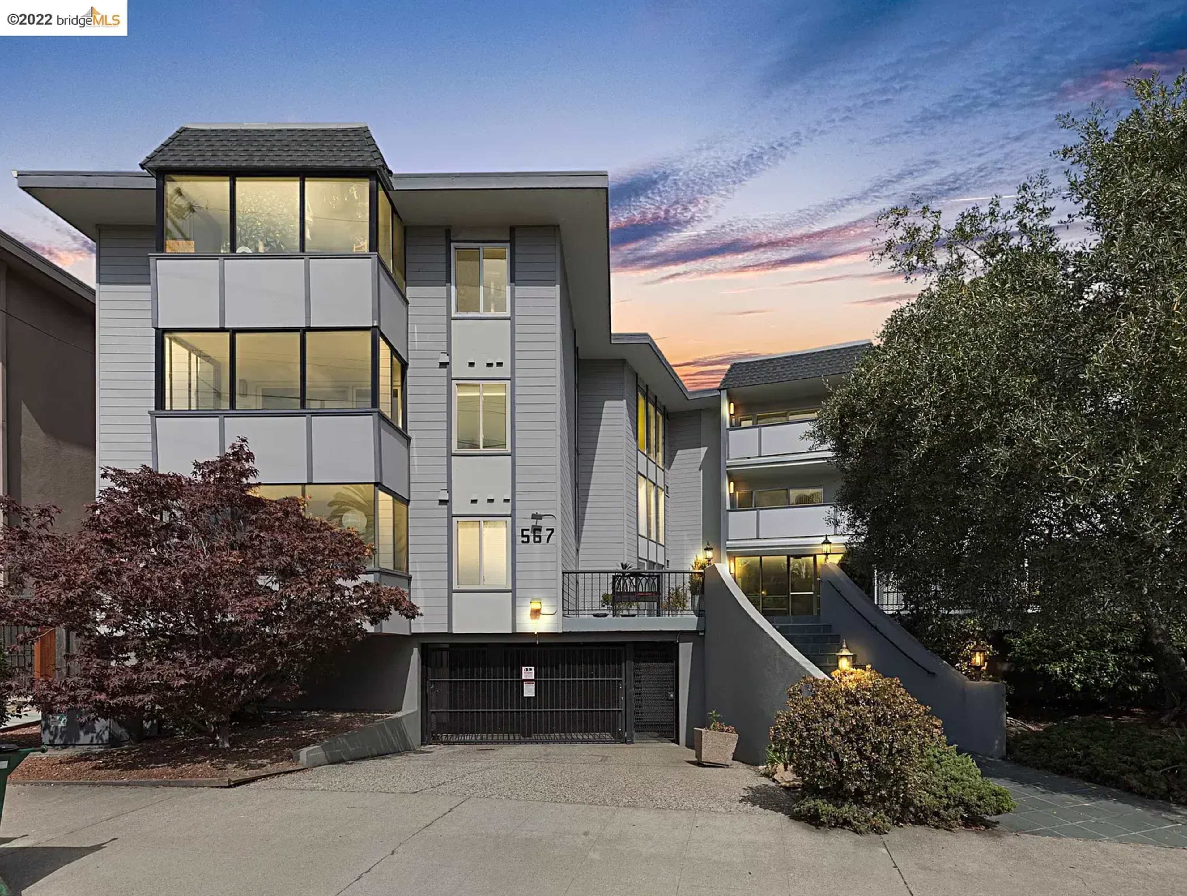 The image shows a three-story modern building with multiple large windows and a driveway leading to a parking area.