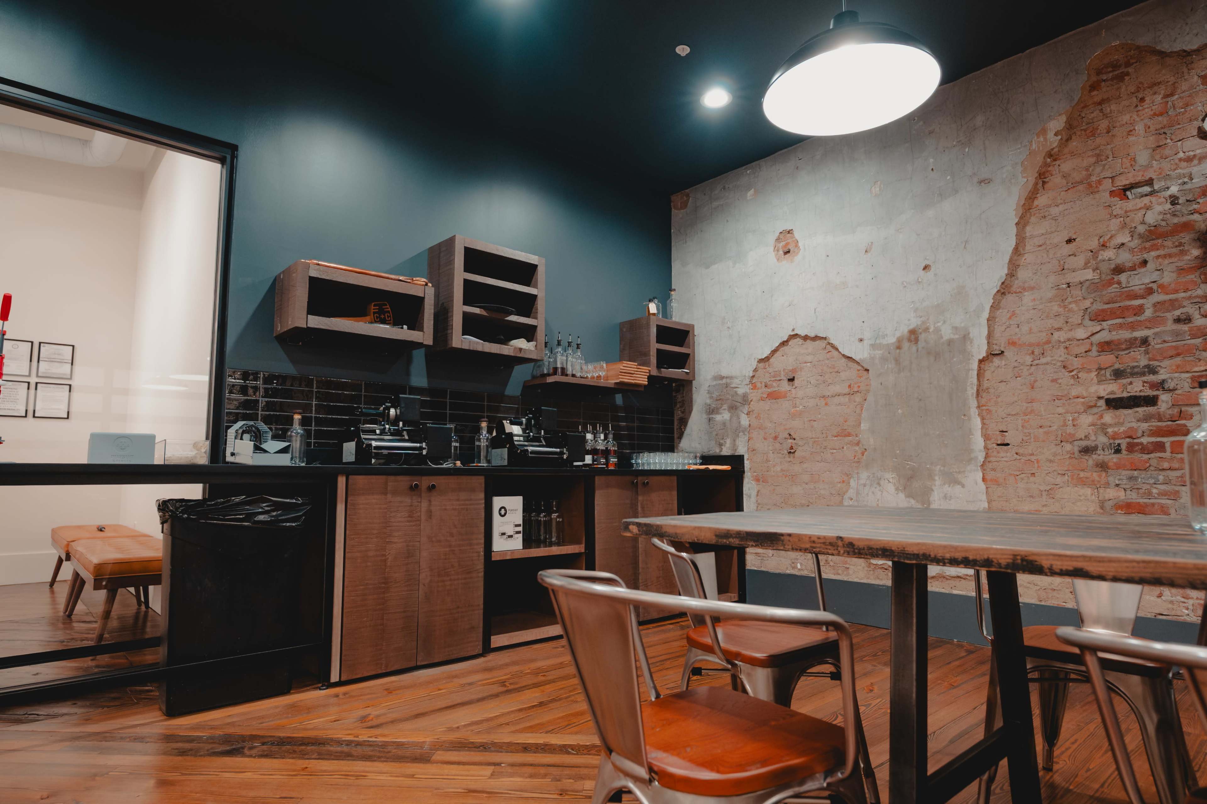 The image features a modern kitchen area with exposed brick walls, wooden cabinetry, and a large table surrounded by metal chairs.