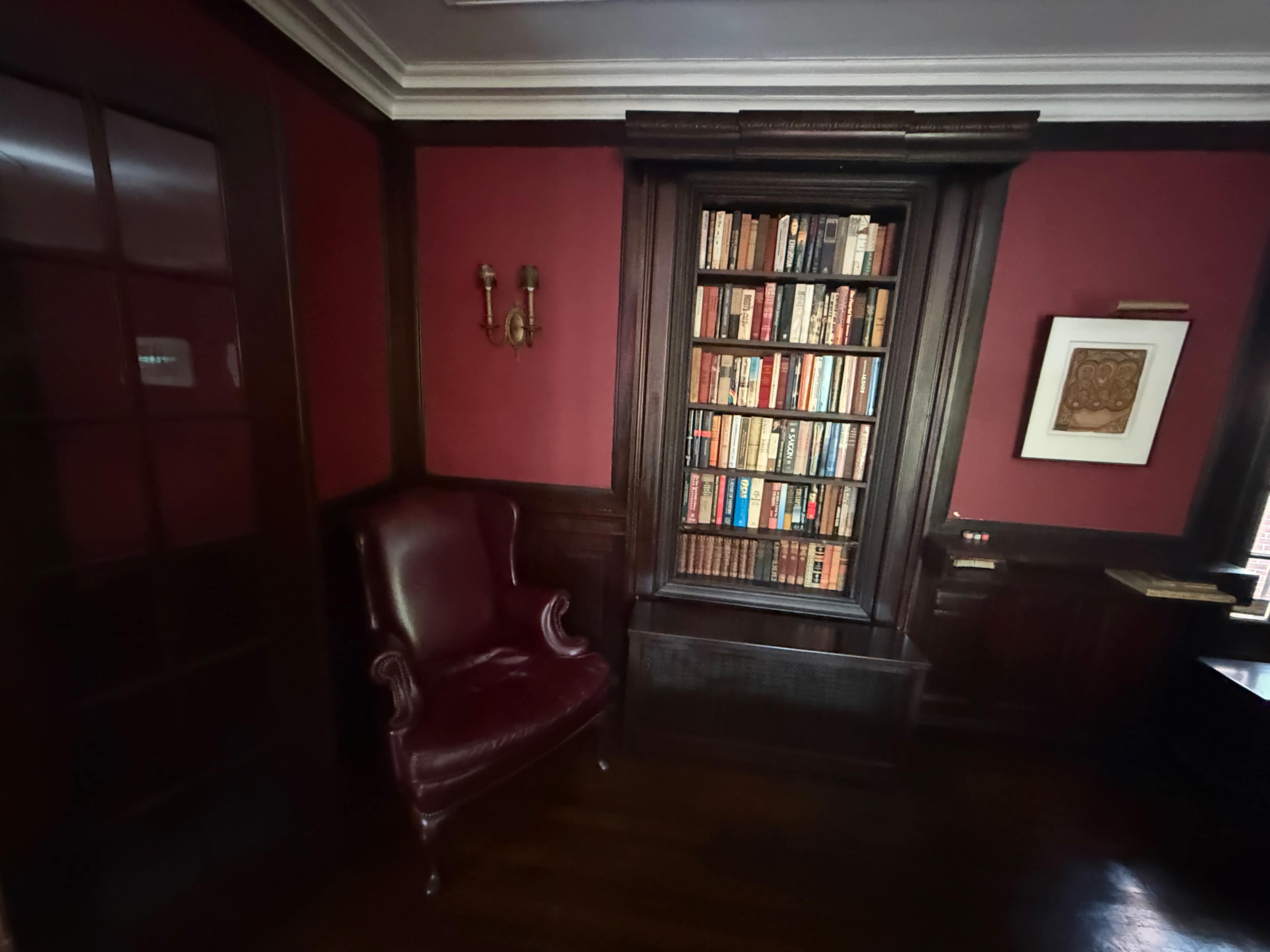 The image shows a dimly lit room with dark red walls, featuring a wooden bookshelf filled with books and a burgundy armchair in the corner.