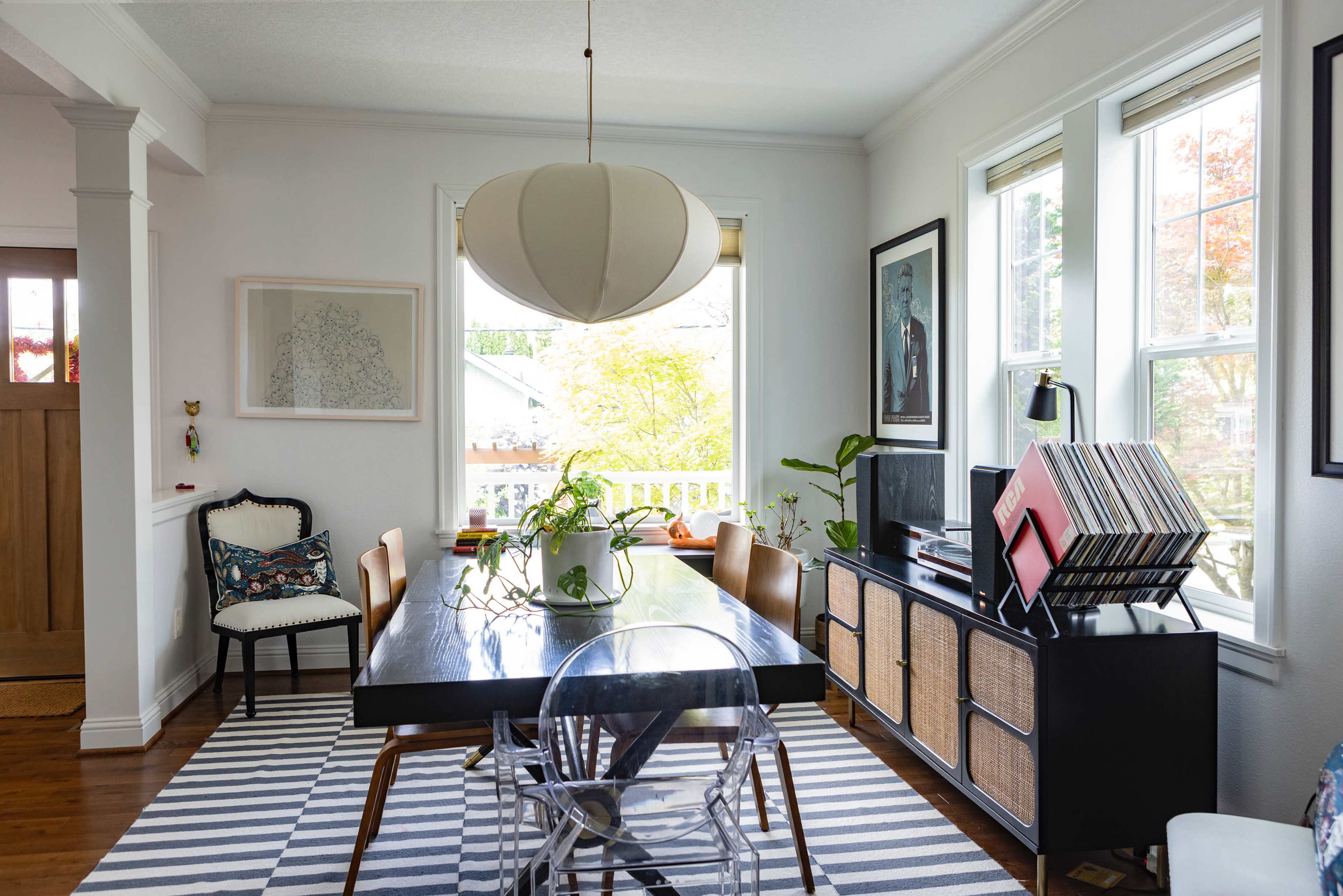 The image shows a dining area featuring a dark wooden table surrounded by chairs, a modern light fixture overhead, and large windows allowing natural light to enter the space.
