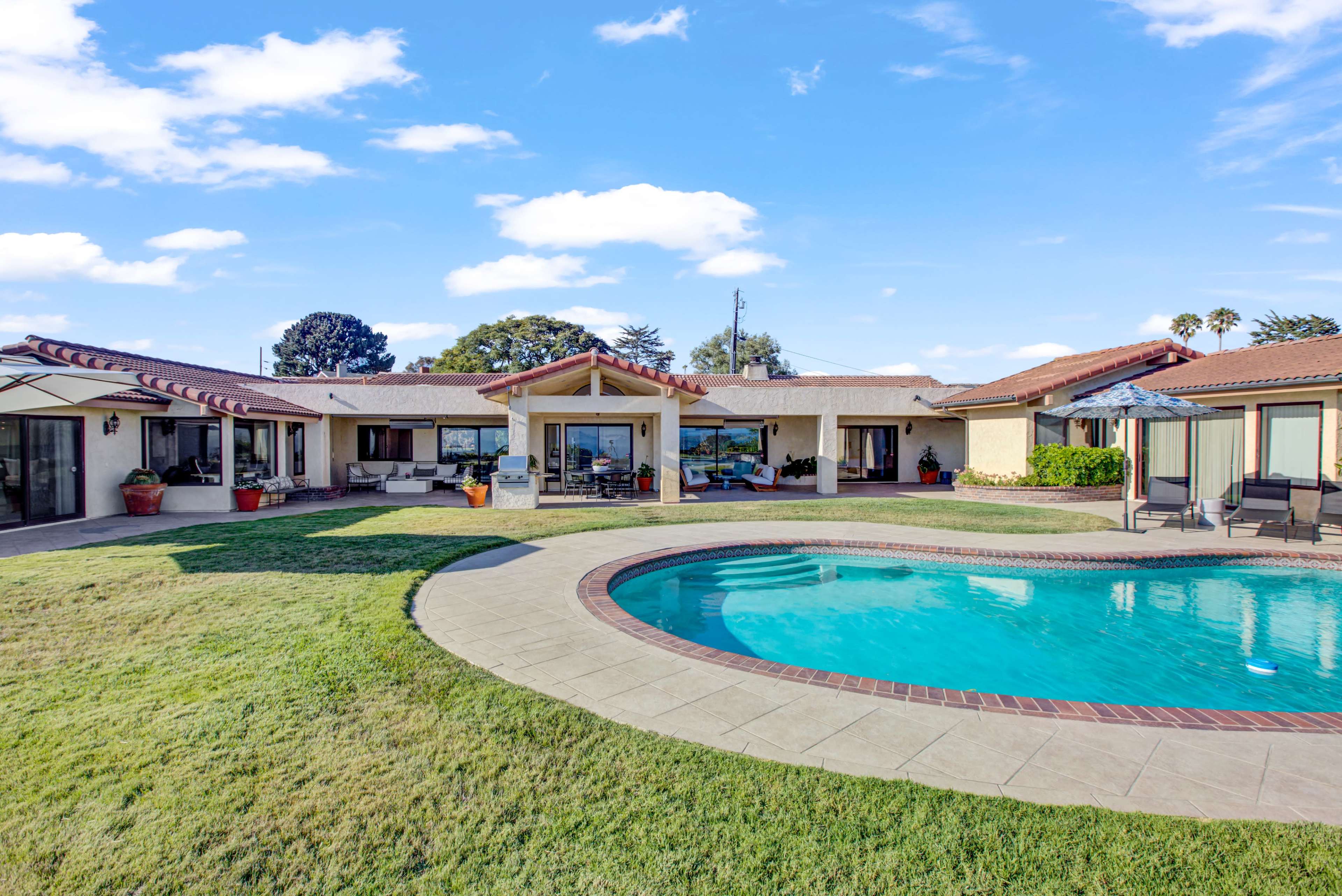 A single-story house with a curved swimming pool in the foreground and a well-maintained lawn surrounded by multiple outdoor seating areas.