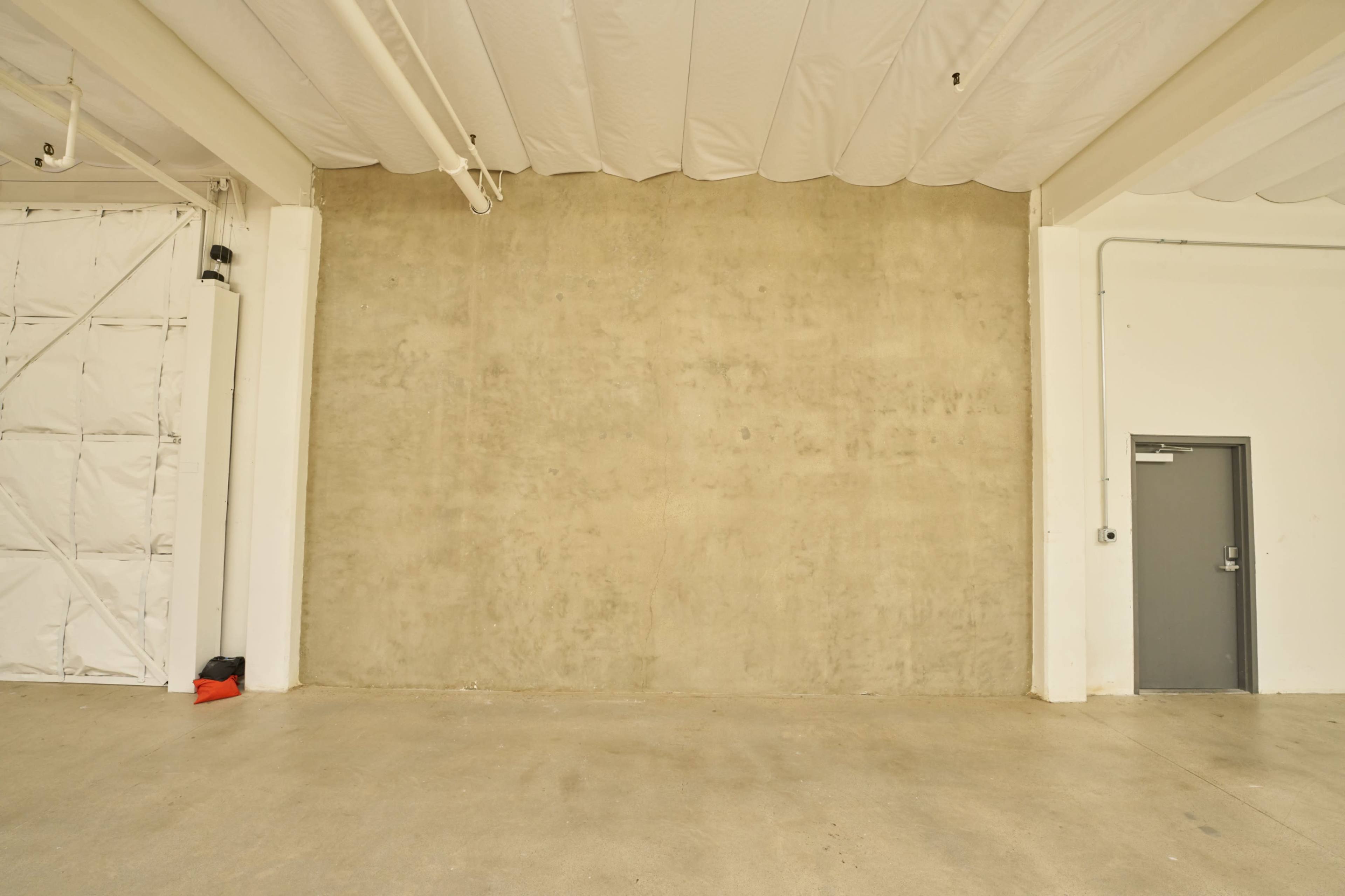 The image shows a large, empty concrete wall in an industrial space with a gray door on the right.