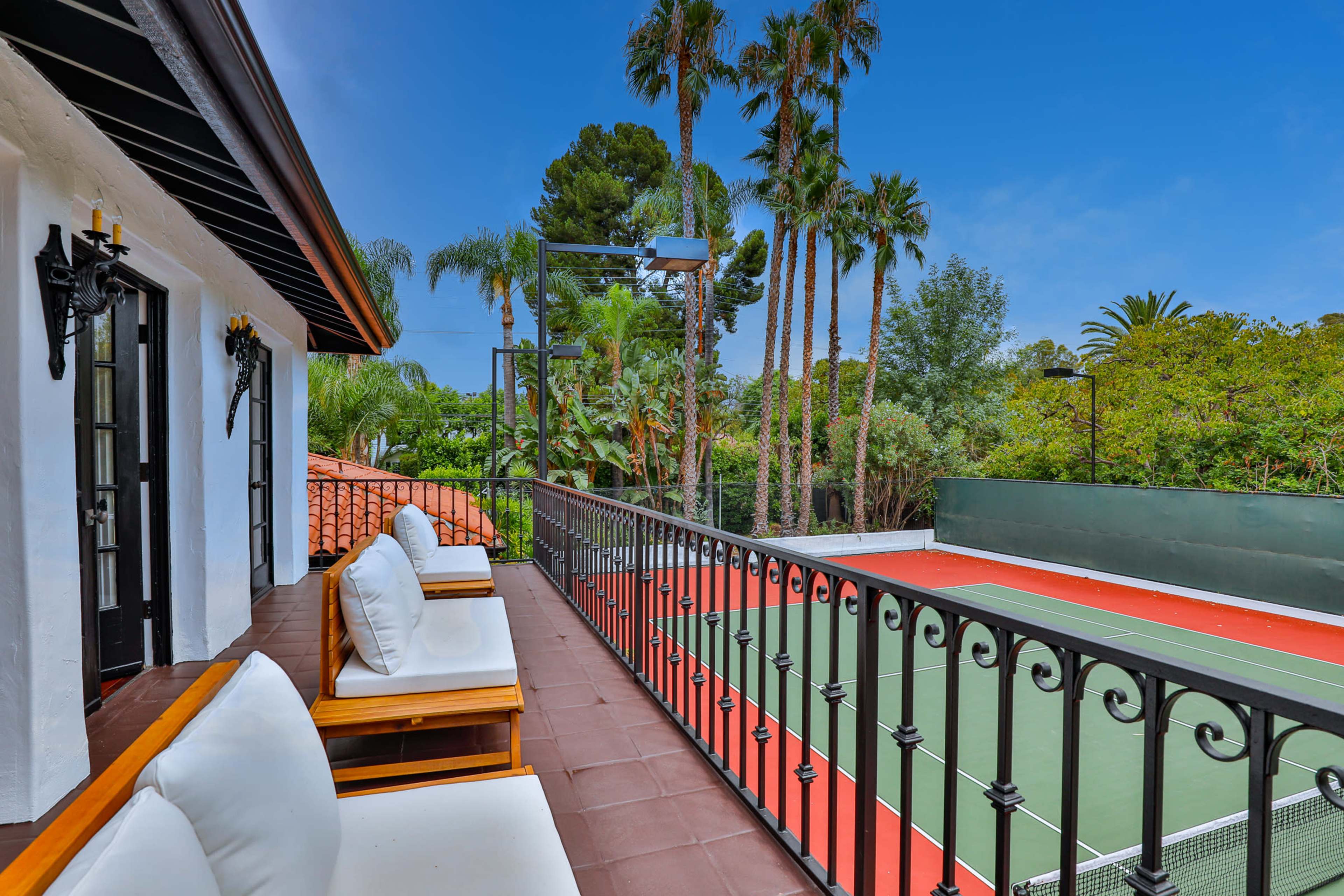 A balcony with white seating overlooks a green tennis court surrounded by palm trees and lush vegetation.