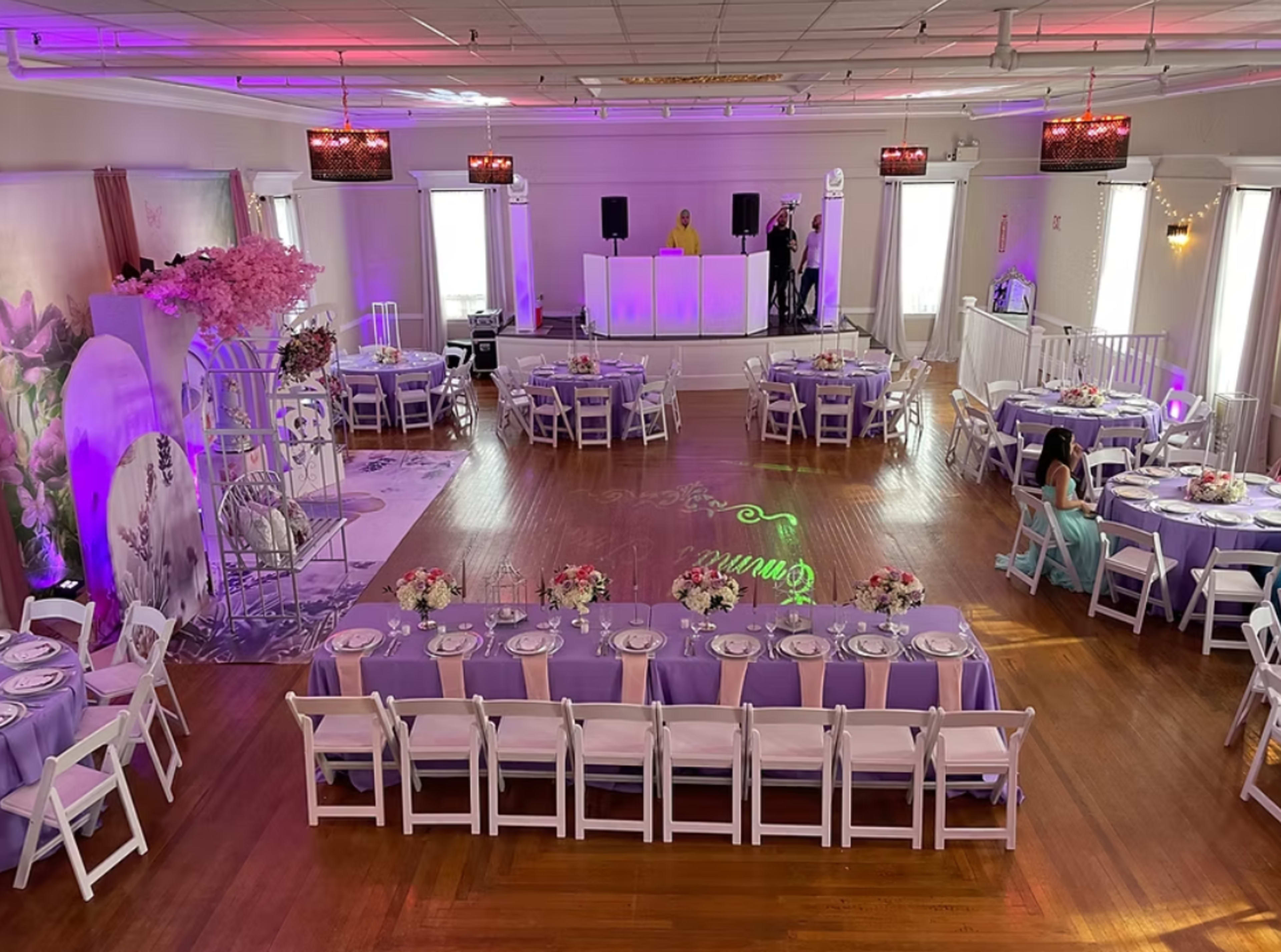The image shows a spacious event hall set up for a celebration, featuring round tables with lavender tablecloths and floral centerpieces, a long rectangular table at the front, and a DJ booth with speakers in the background.
