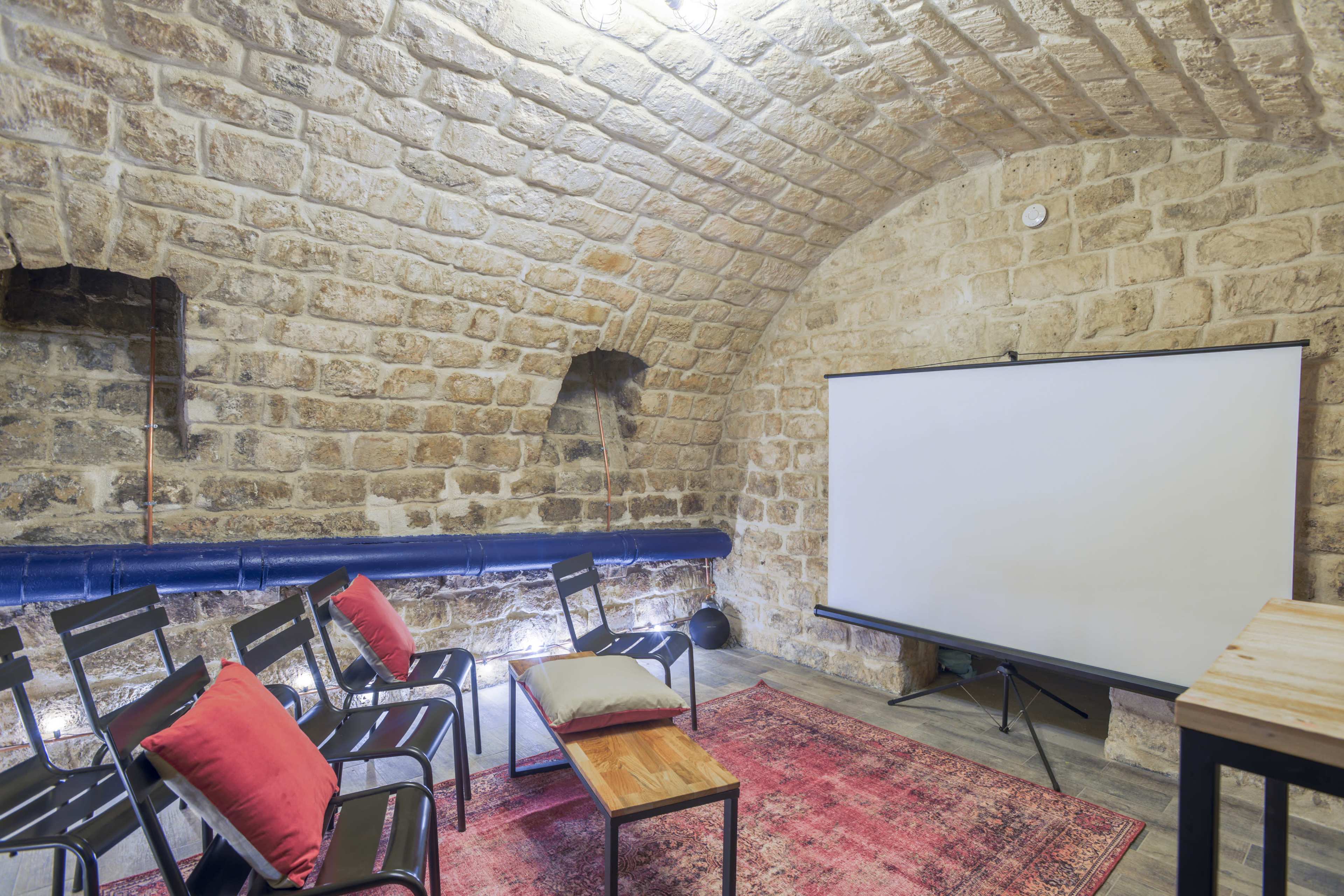 A rustic stone room with a curved ceiling, featuring several black chairs, a coffee table, and a large projection screen.