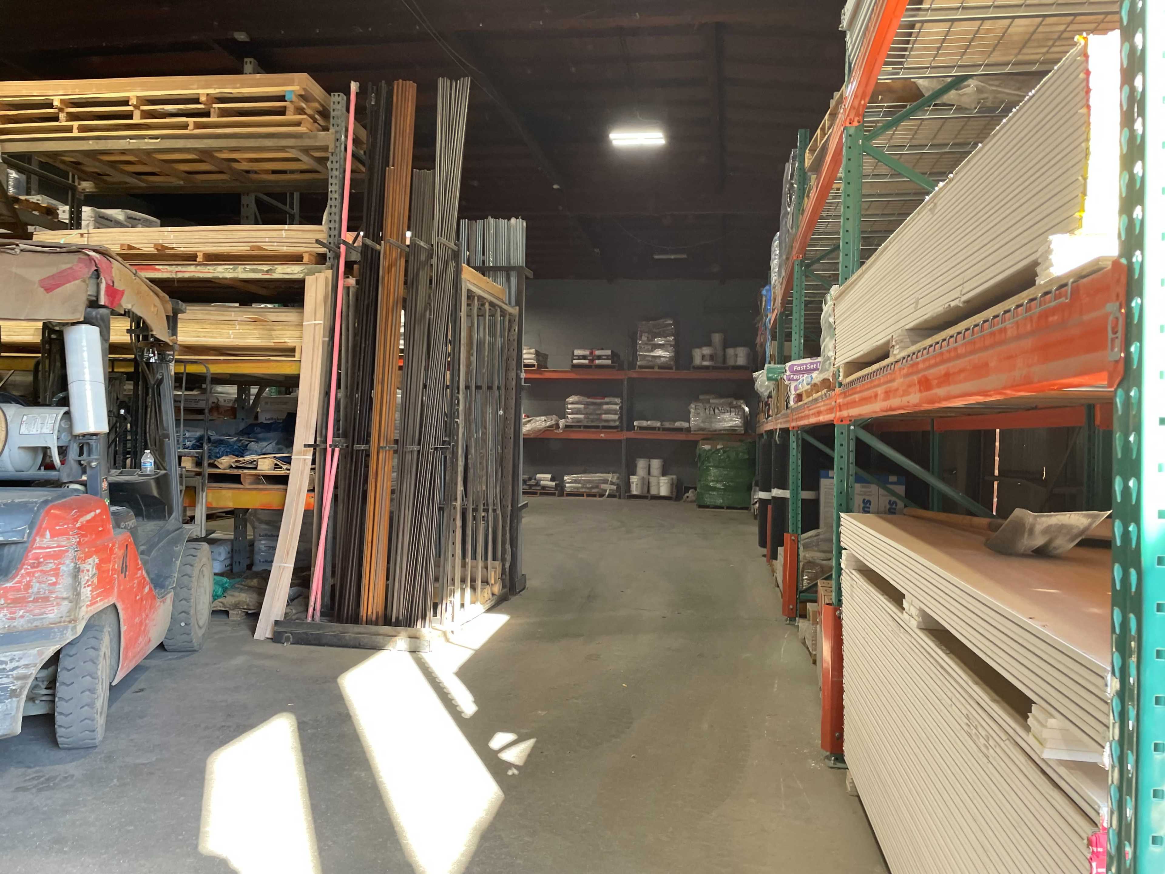 The image shows a warehouse aisle lined with shelves holding building materials and a forklift parked on the left.