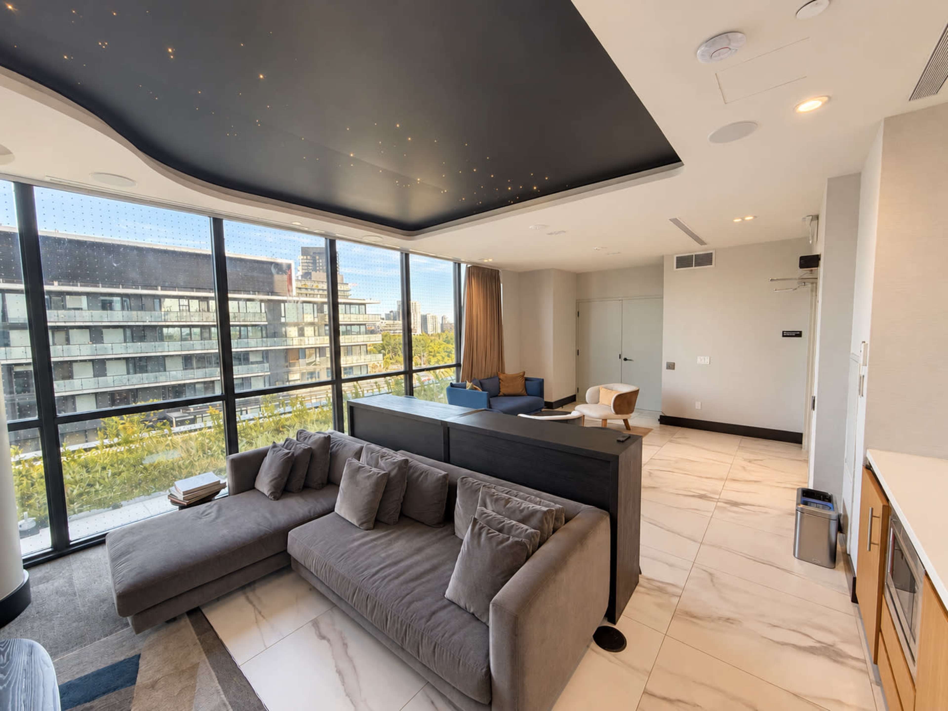 A modern living space features a gray sectional sofa, a sleek kitchen area, and large windows providing a view of the outdoors.