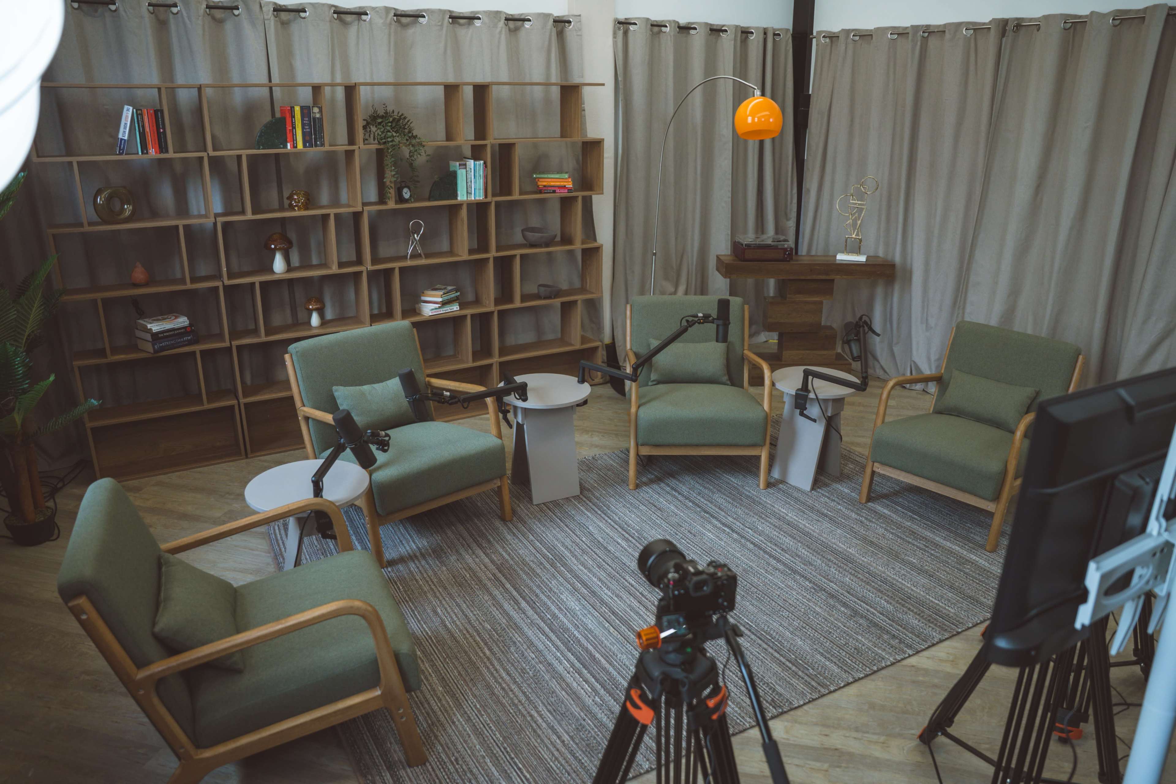 The image shows a well-arranged interview setup with four green chairs, a small table, and a bookshelf, all positioned in a room with gray curtains and a warm light fixture.