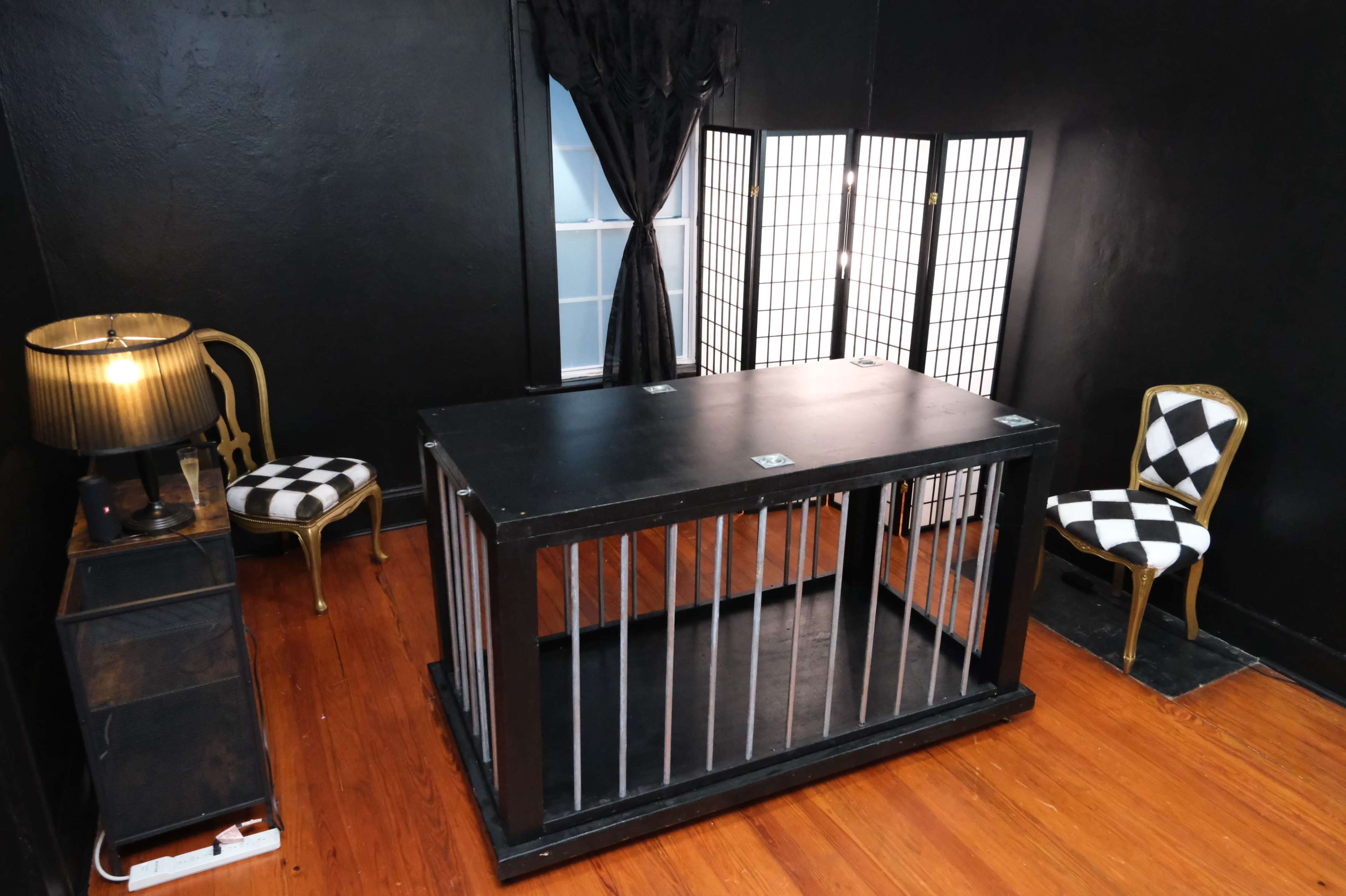 The image shows a black-walled room featuring a large black table with metal bars, a lamp, a patterned chair, and a folding screen.