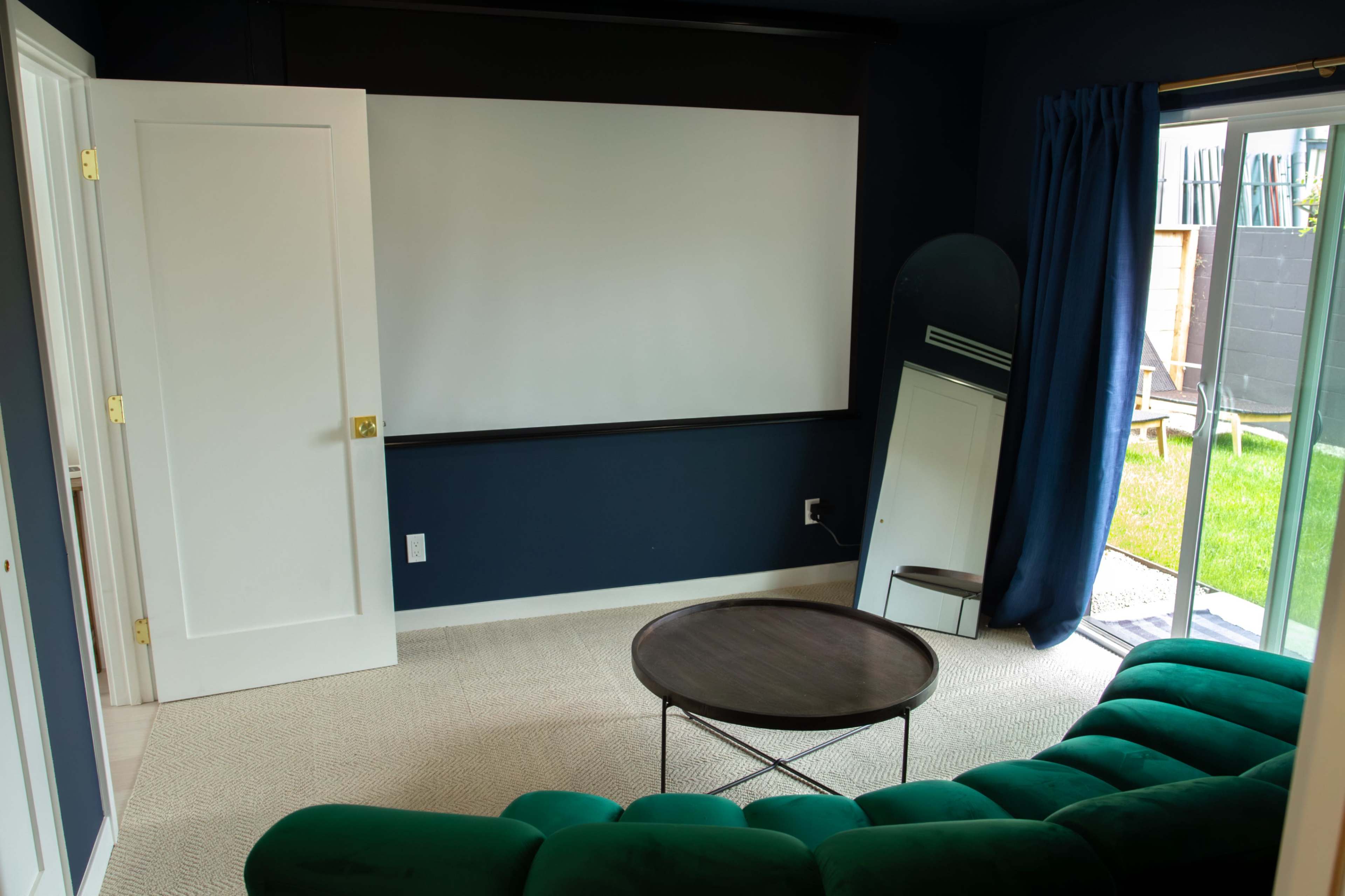 The image shows a small living room with a green sectional sofa, a round coffee table, a blank wall suitable for a projector, and a sliding glass door leading to a grass area outside.