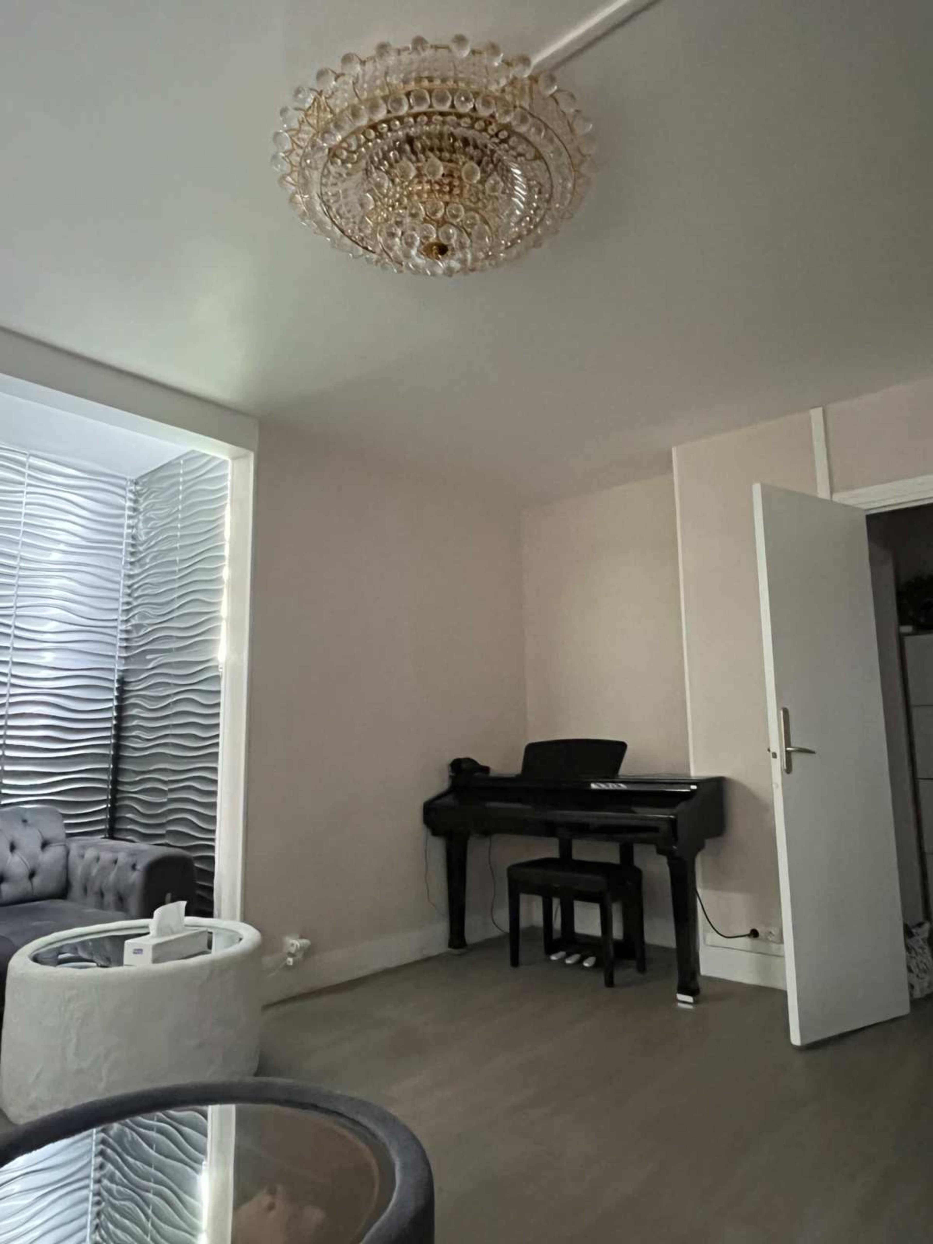 The image shows a modern room featuring a black piano, a chandelier, and a sofa set, with light-colored walls and flooring.