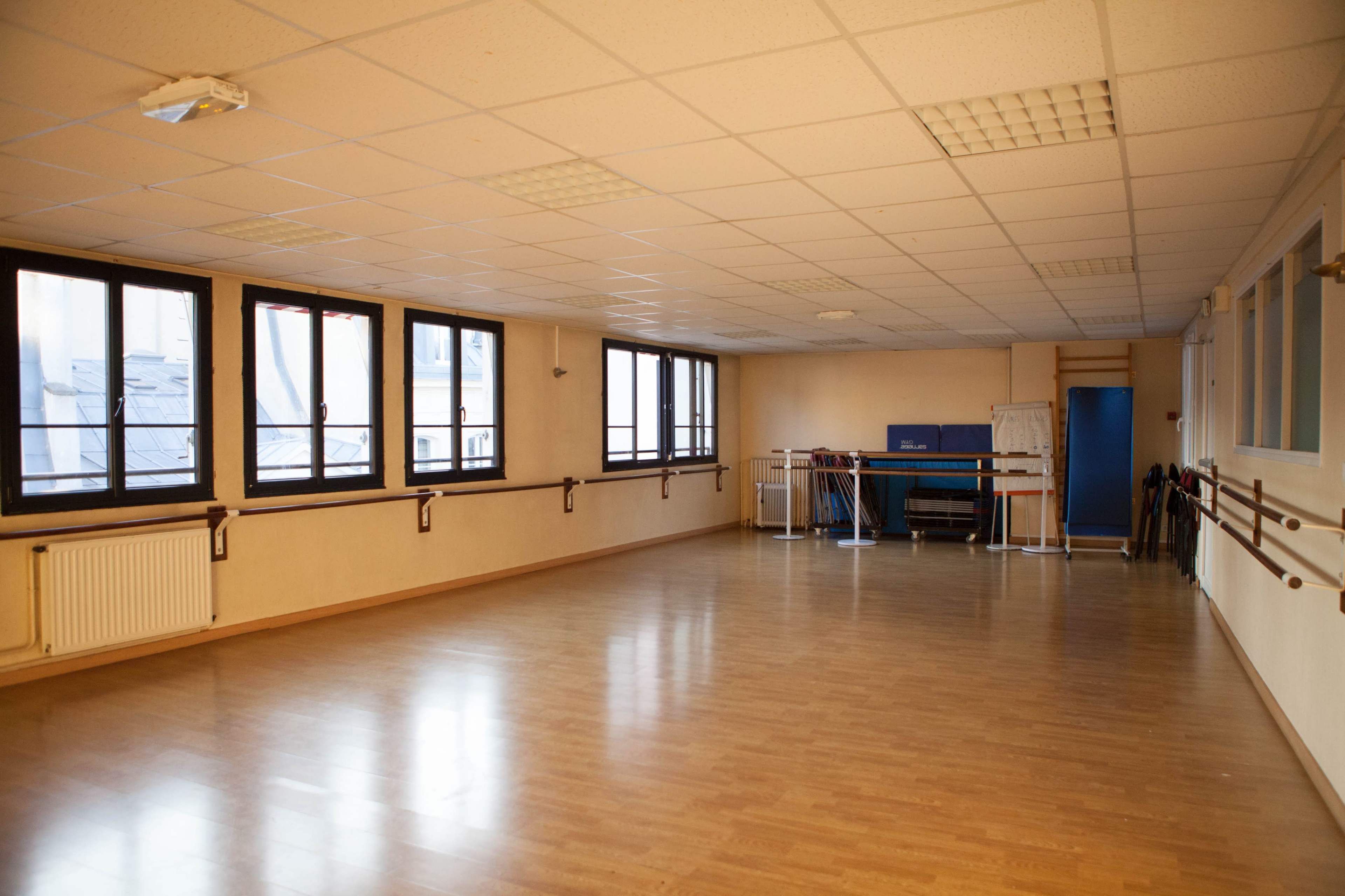 The image shows an empty dance studio with wooden flooring, large windows, and mirrors along one wall.