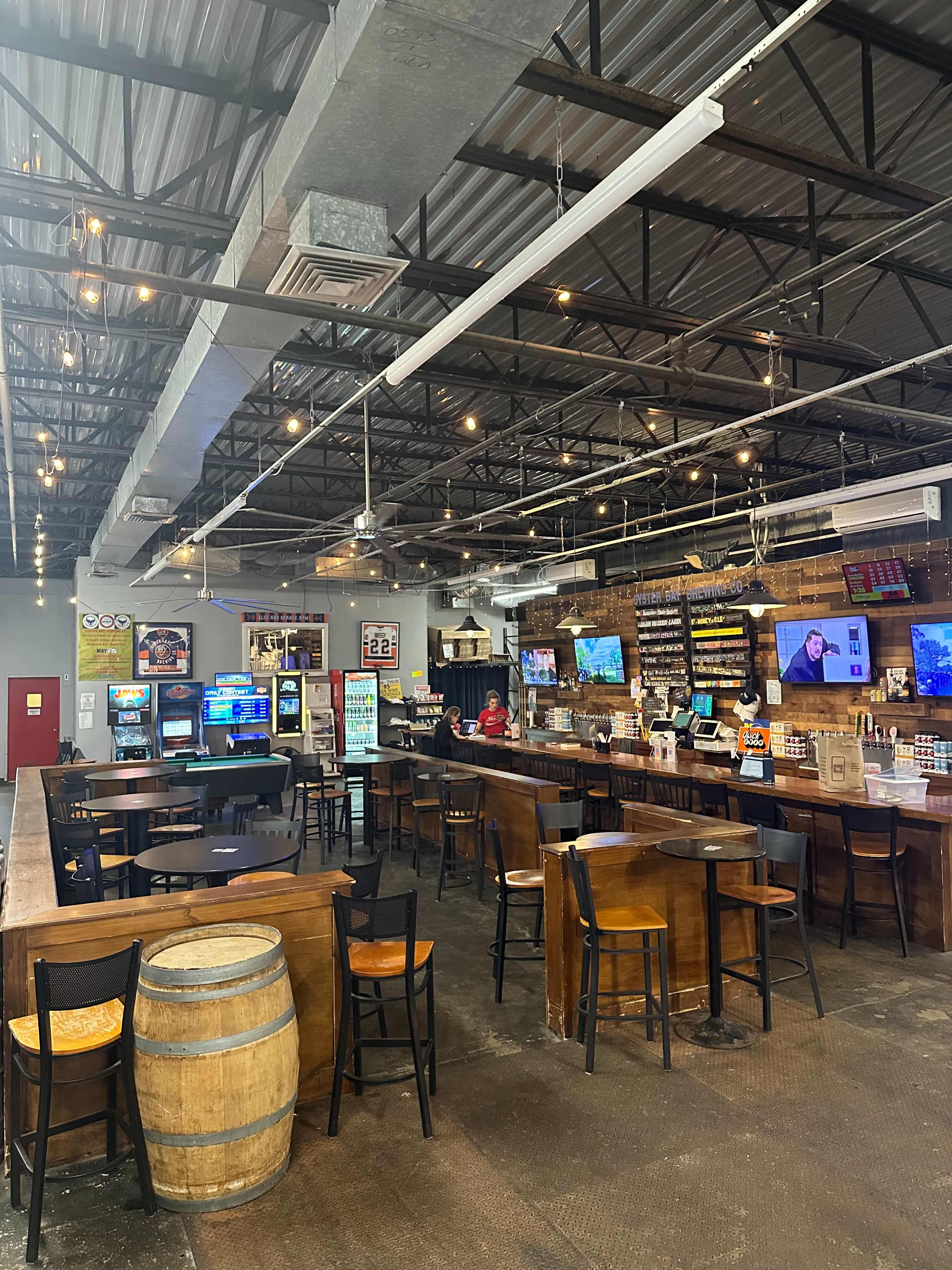 The image shows a spacious bar area with wooden furniture, multiple screens displaying content, and a long countertop serving as the bar, illuminated by overhead lights.