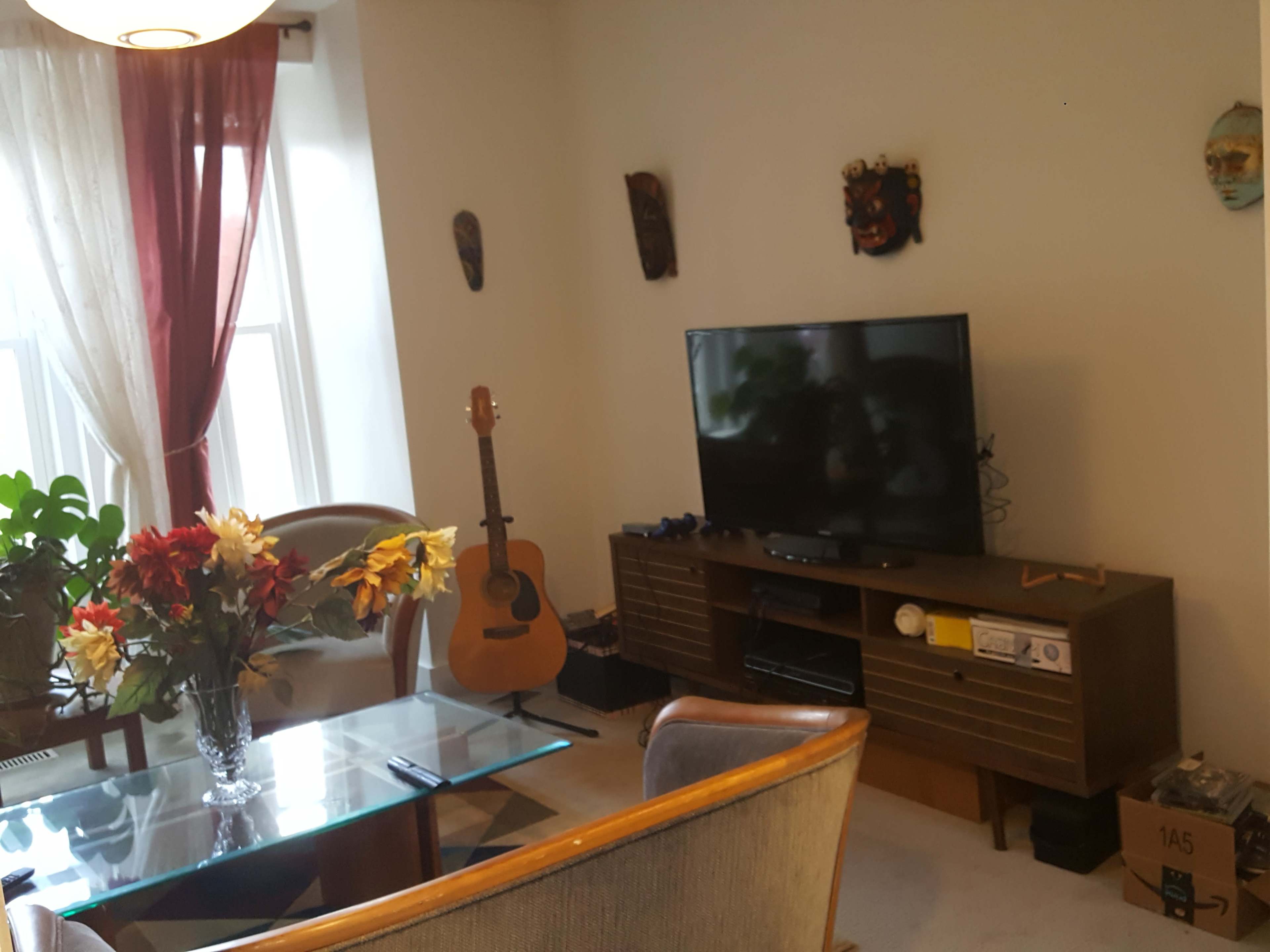 A living room with a glass dining table, a TV on a wooden stand, a guitar leaning against the wall, and decorative masks hanging above.