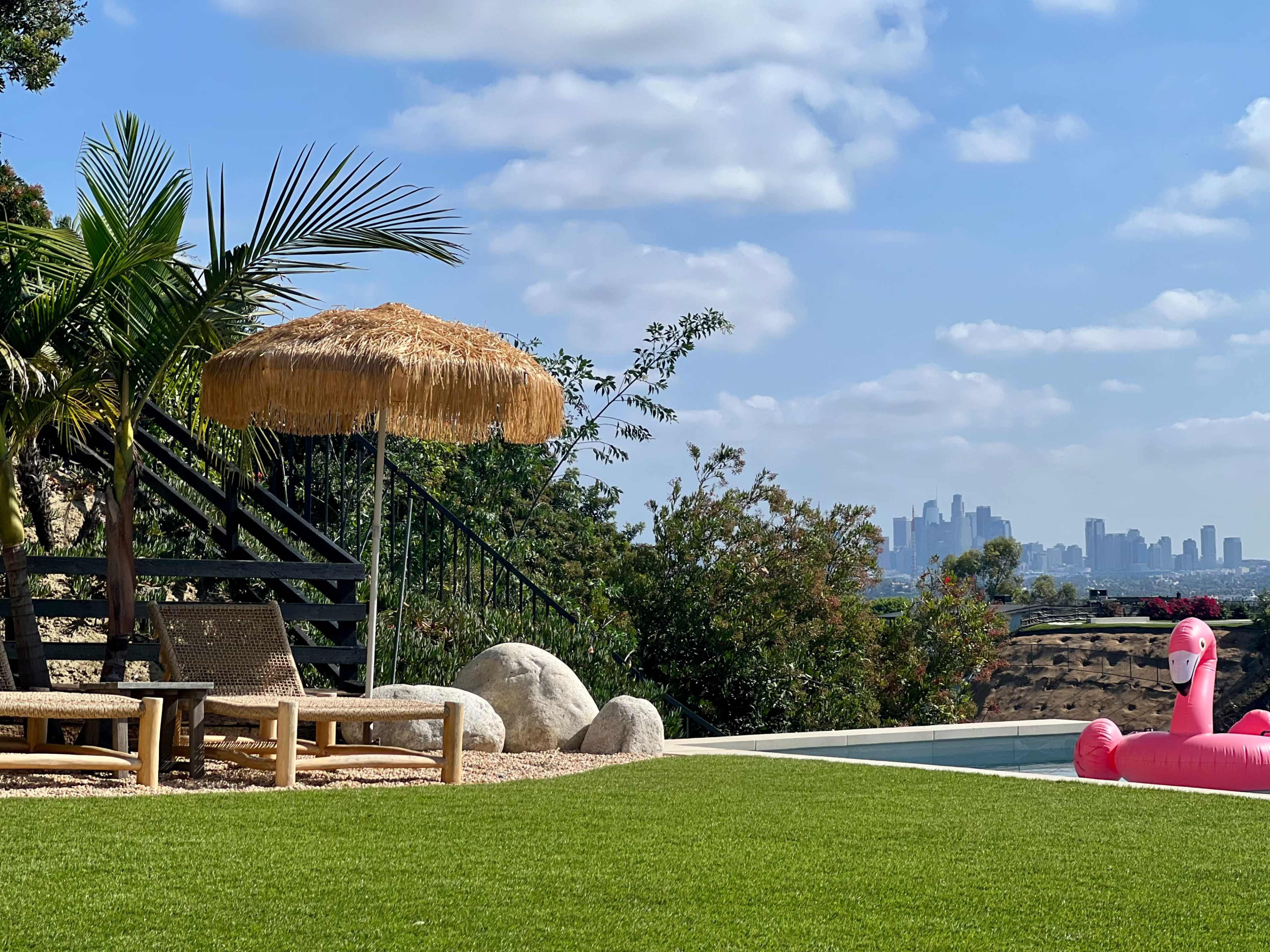 A straw umbrella stands beside lounge chairs on a grassy area overlooking a swimming pool and the skyline of a city in the background.